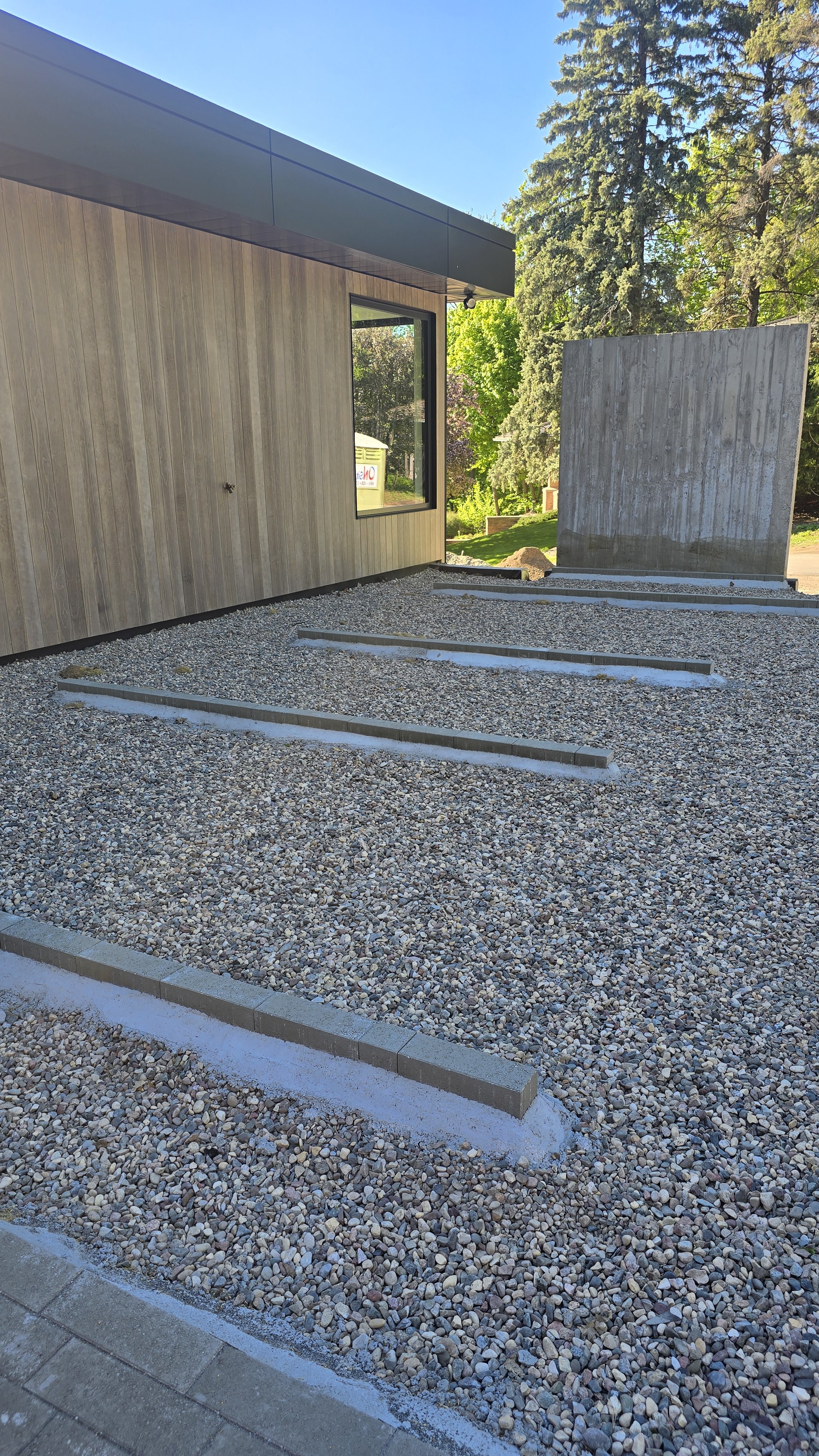Outdoor construction scene with a modern building featuring wood siding and large window, gravel paving with inset lines of pavers, a board-formed concrete wall, and tall trees in the background under a clear blue sky.