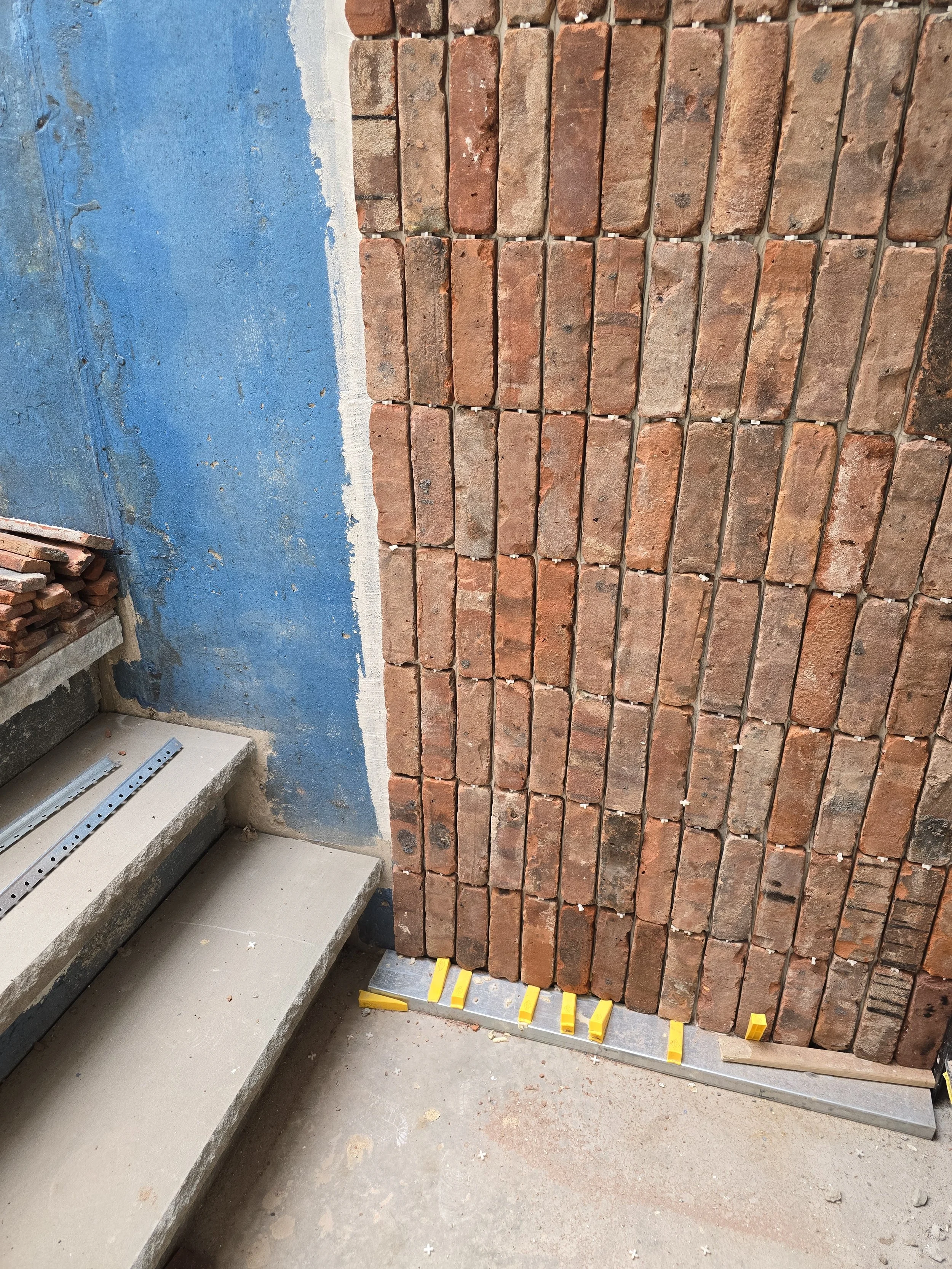 Exterior of a building under construction with a reclaimed brick veneer wall, unfinished concrete stairs, and construction tools and materials on the floor.