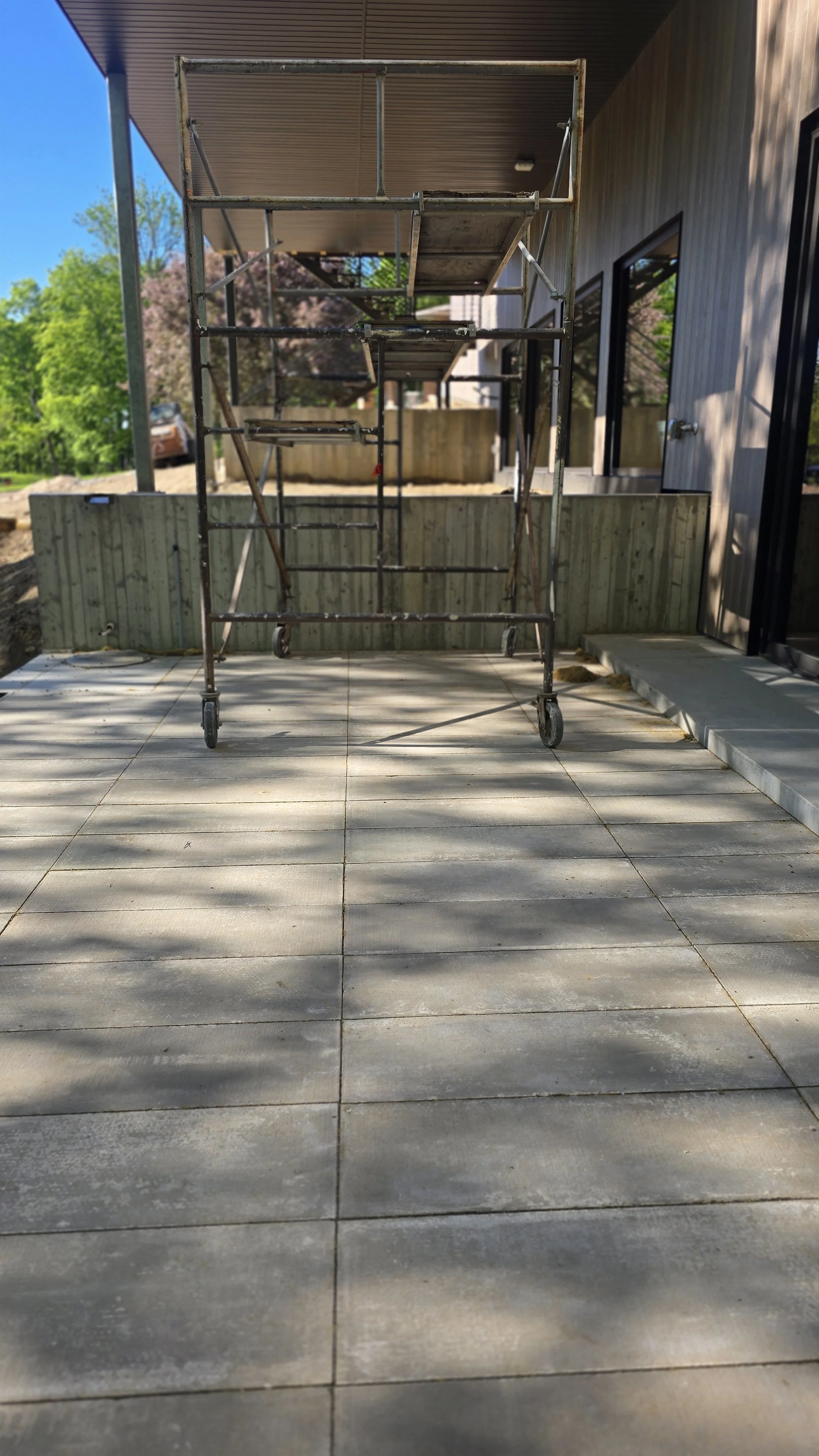 Scaffolding set up on a patio in front of a house with a grey wooden exterior. Shadows cast by nearby trees are visible on the tiled ground.
