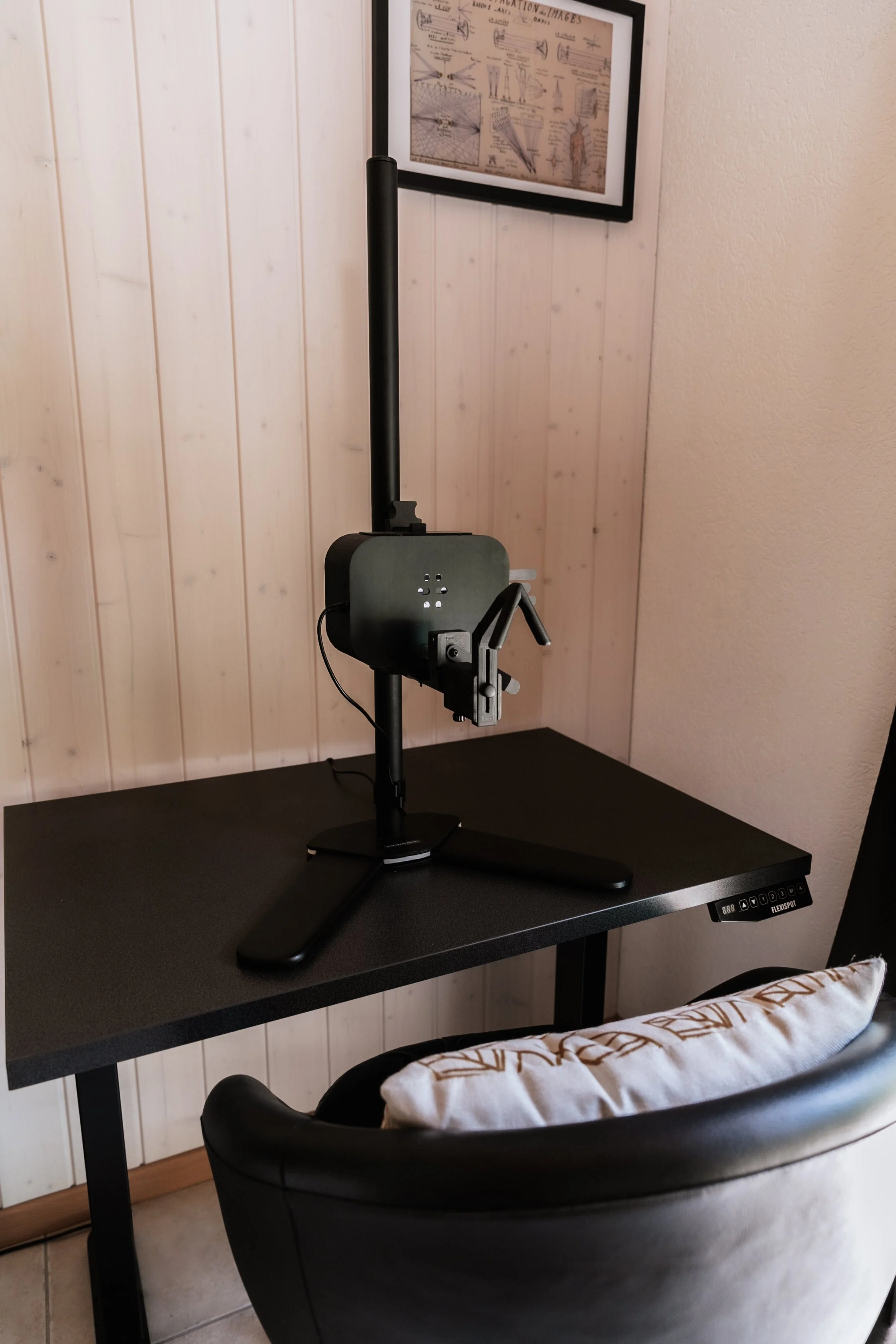 Un appareil de simulation de tir ou de tir à l'arc monté sur un bureau noir, avec un fauteuil en avant et un cadre avec des schémas dans le fond, dans une pièce avec un mur en bois clair.
