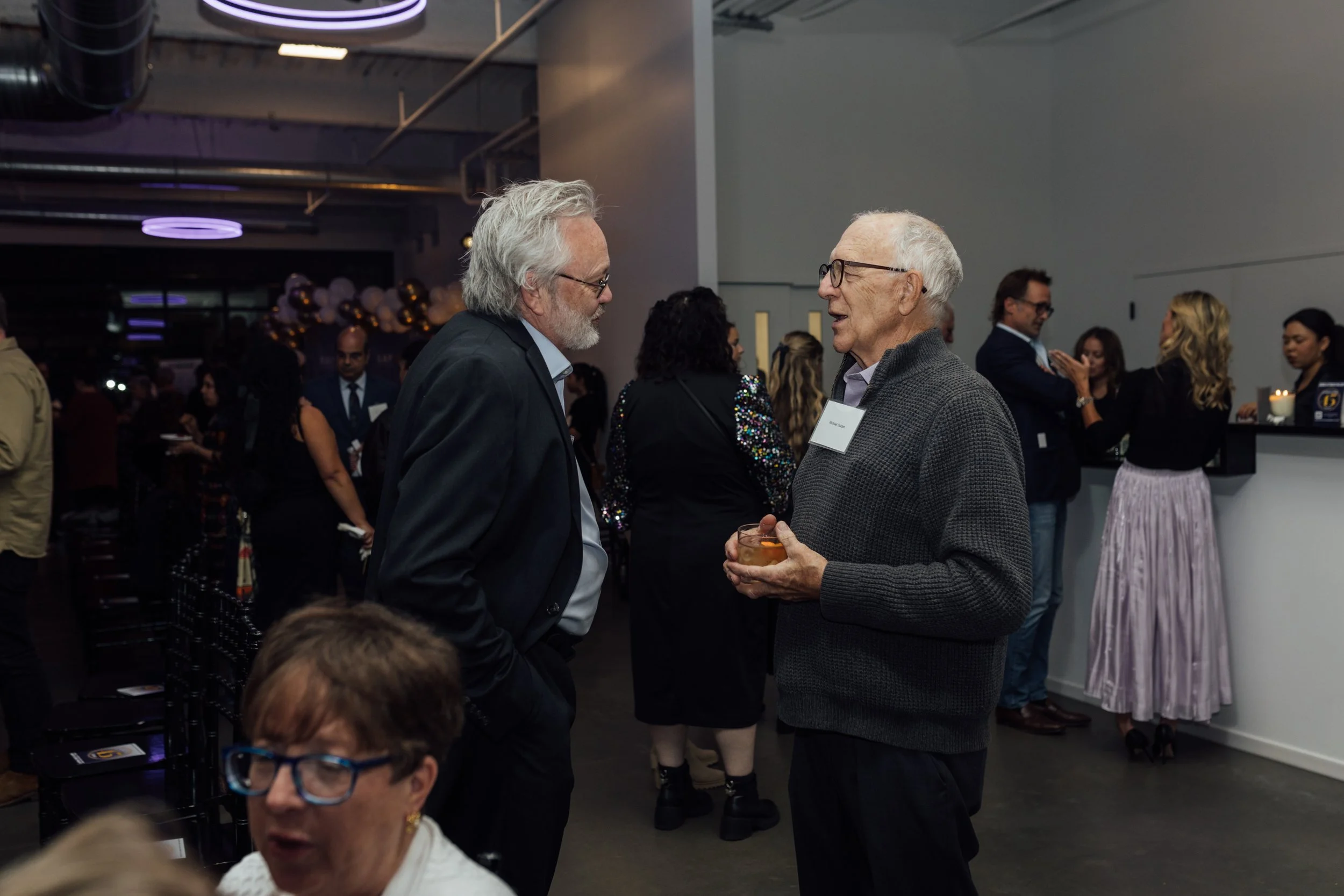 Two elderly men are talking at a social event, one wearing a suit and glasses, the other holding a drink and wearing a gray sweater, with other guests in the background.
