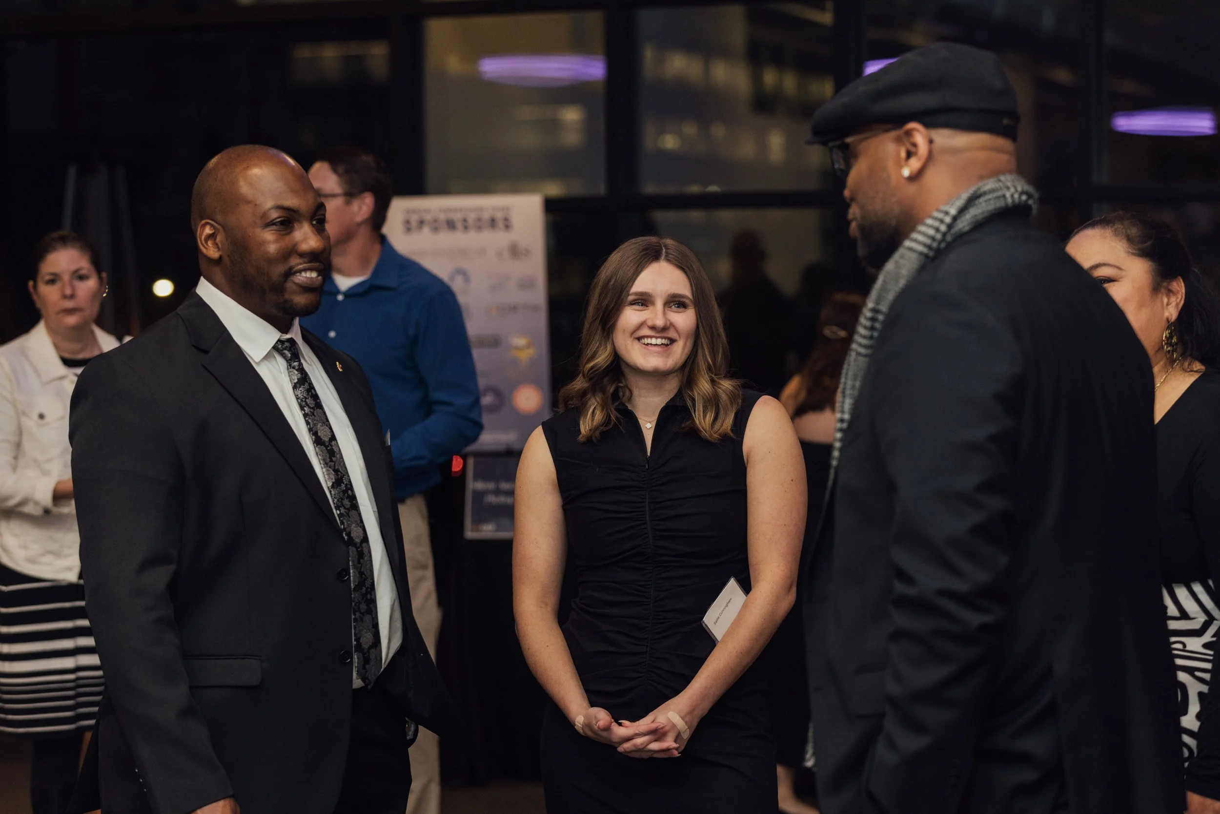 A group of people engaged in conversation at an indoor event, with a woman smiling in the center, wearing a black dress, and men on either side dressed in suits and dark clothing.