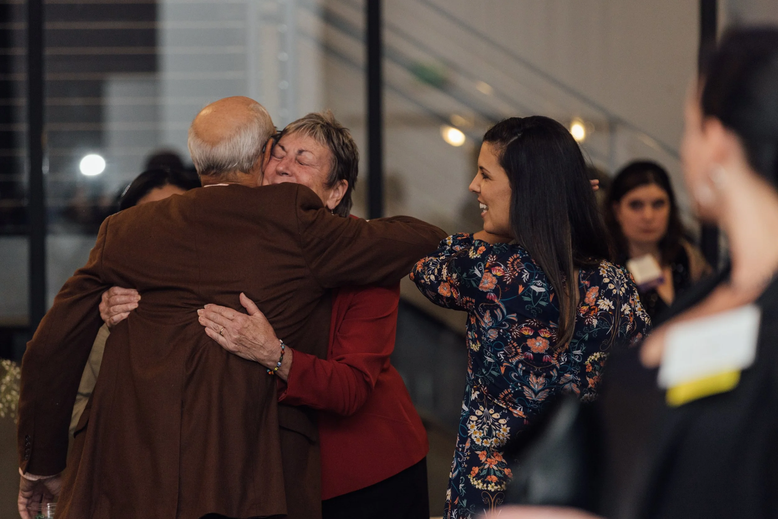 An elderly man and woman hugging, while a young woman smiling and looking at them, at a social gathering or celebration.