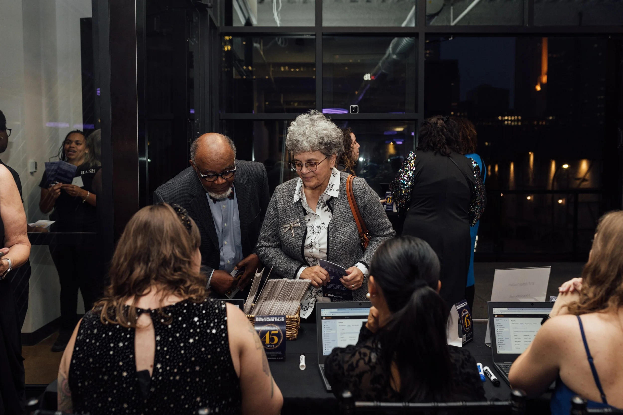 People gathered around a table at a networking event or conference, engaging in conversation and browsing laptops and brochures in a modern indoor setting at night.