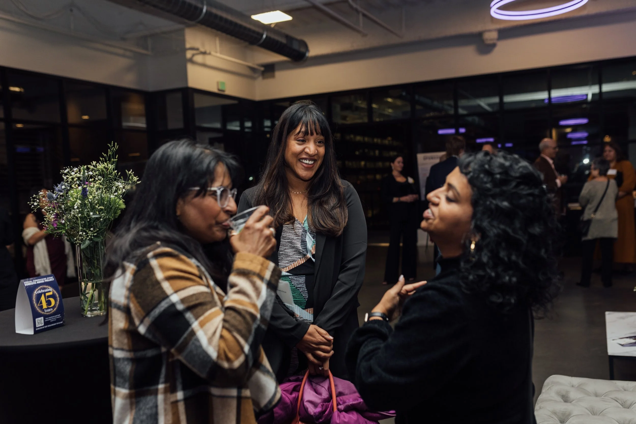 Three women at a social event, one drinking from a glass and the others engaged in conversation, with a table of flowers and event signage in the background.