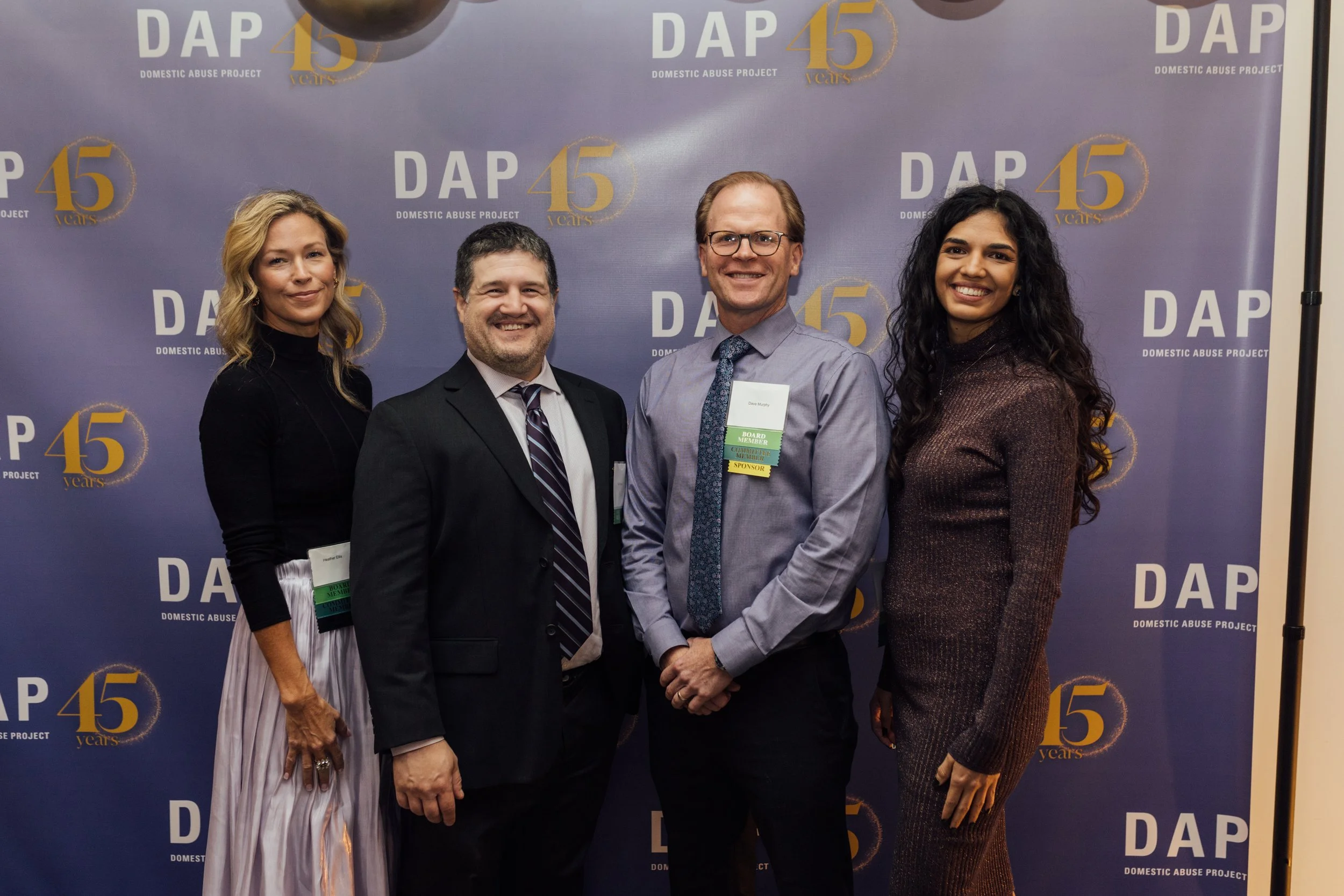 Four people standing in front of a blue backdrop with the logos and text for the Domestic Abuse Project celebrating its 45th anniversary, smiling and posing for the photo.