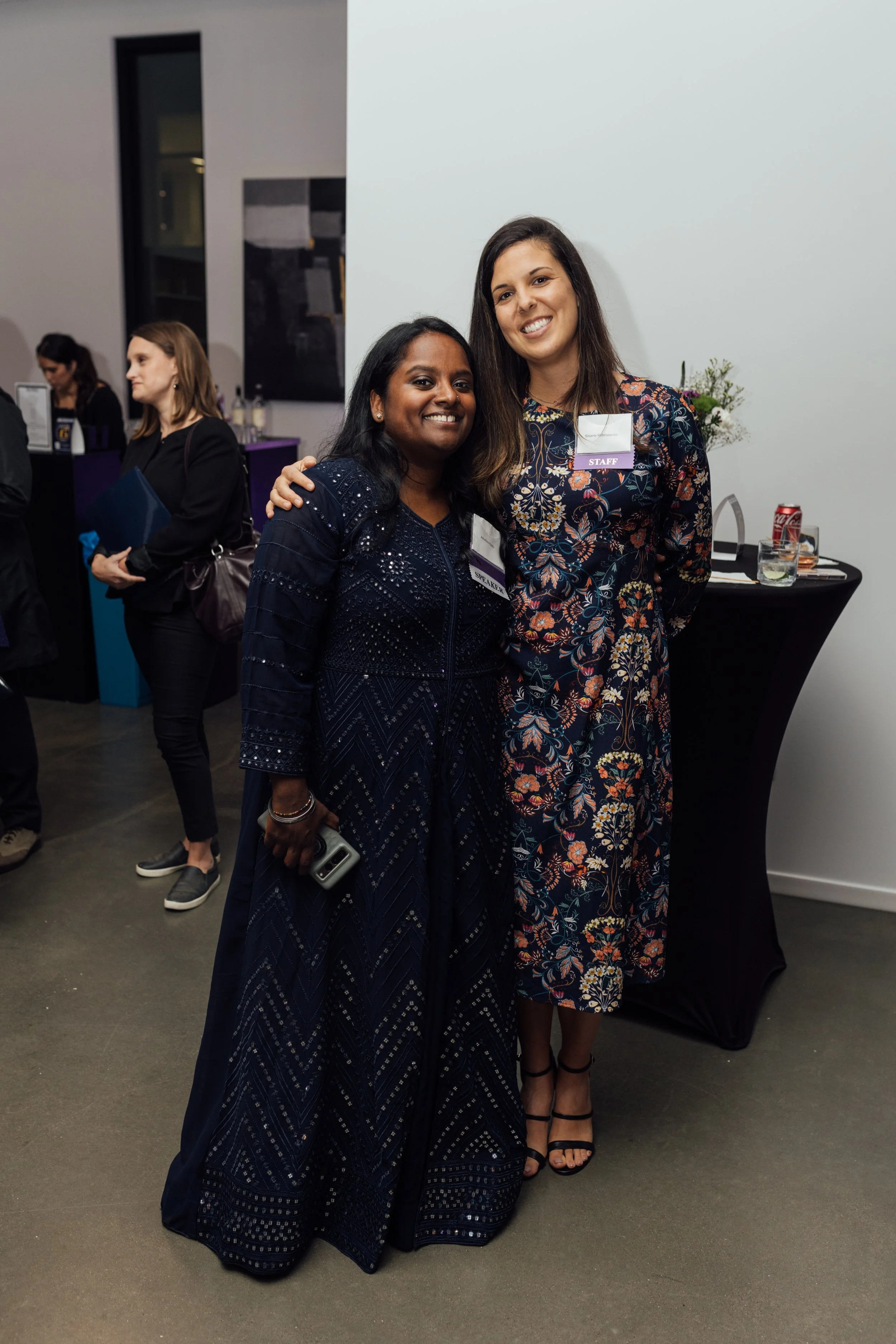 Two women wearing dresses, smiling, and posing for a photo at a semi-formal event, with one woman holding a phone.
