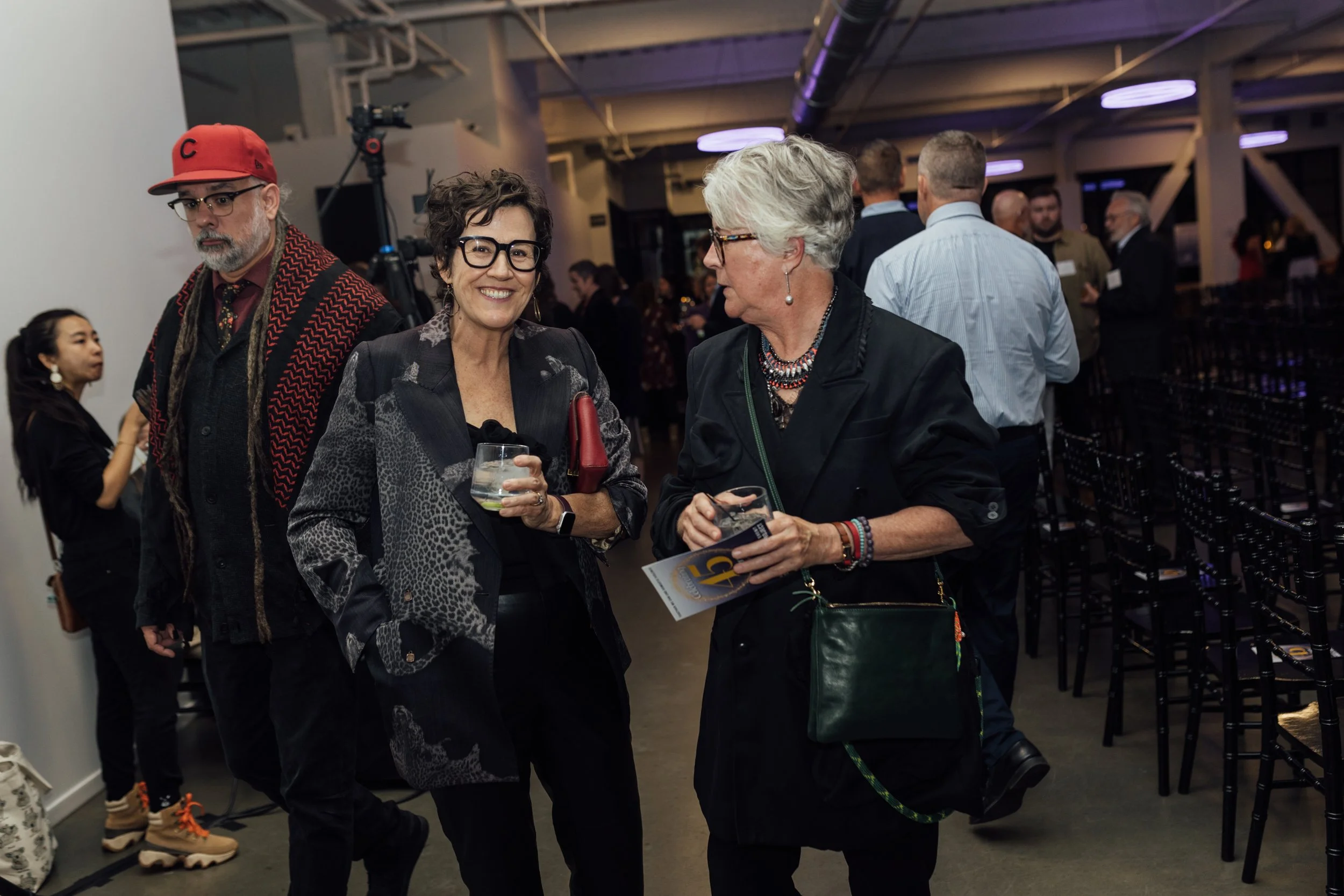 Two women engaged in a conversation holding drinks at a social event, with a man standing nearby and other people mingling in the background.