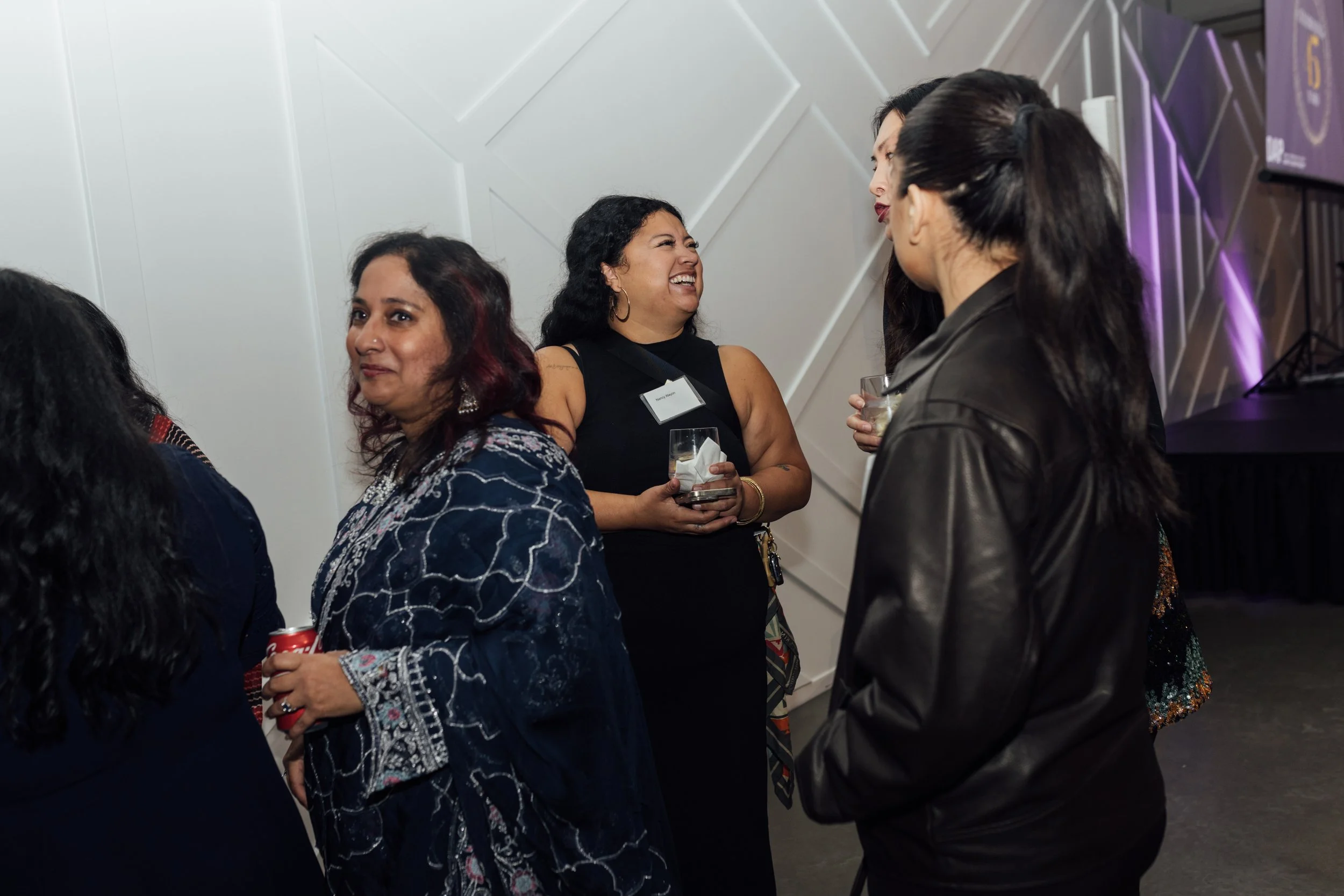 Group of women socializing at a professional event, holding drinks and engaging in conversation in a modern, well-lit venue.