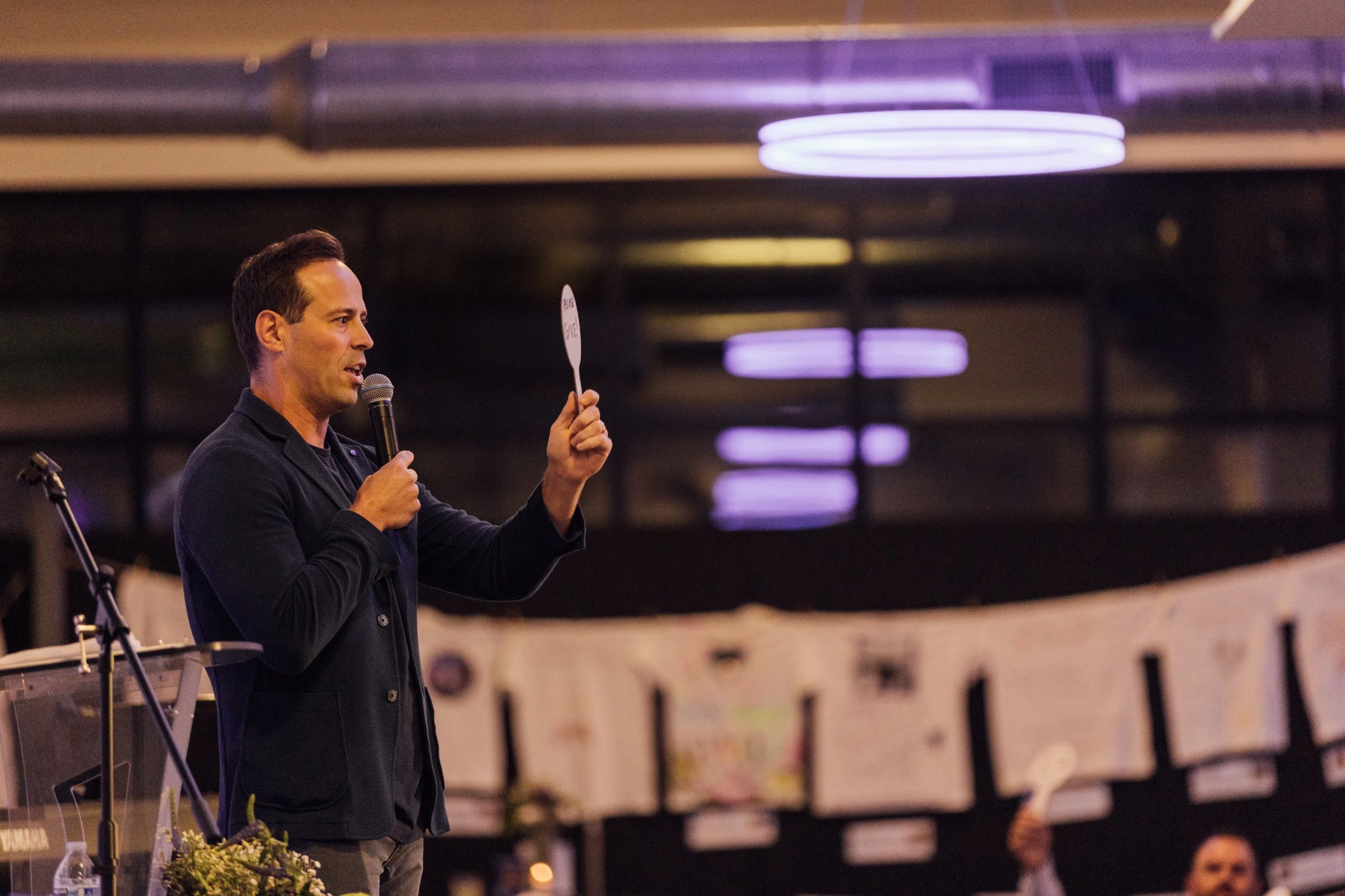 A man holding a microphone and a small white board with writing on it, speaking at an indoor event with purple lighting and banners in the background.