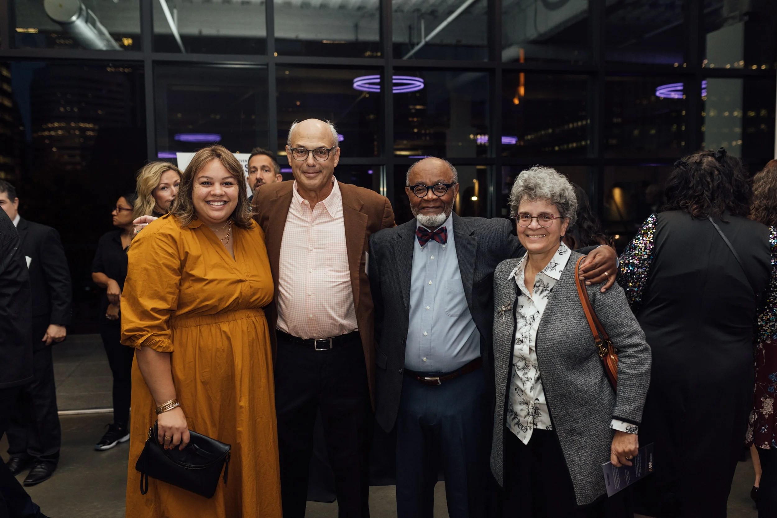 Group of five diverse people smiling and posing together at a social event in a modern indoor venue, with city lights visible through large windows in the background.