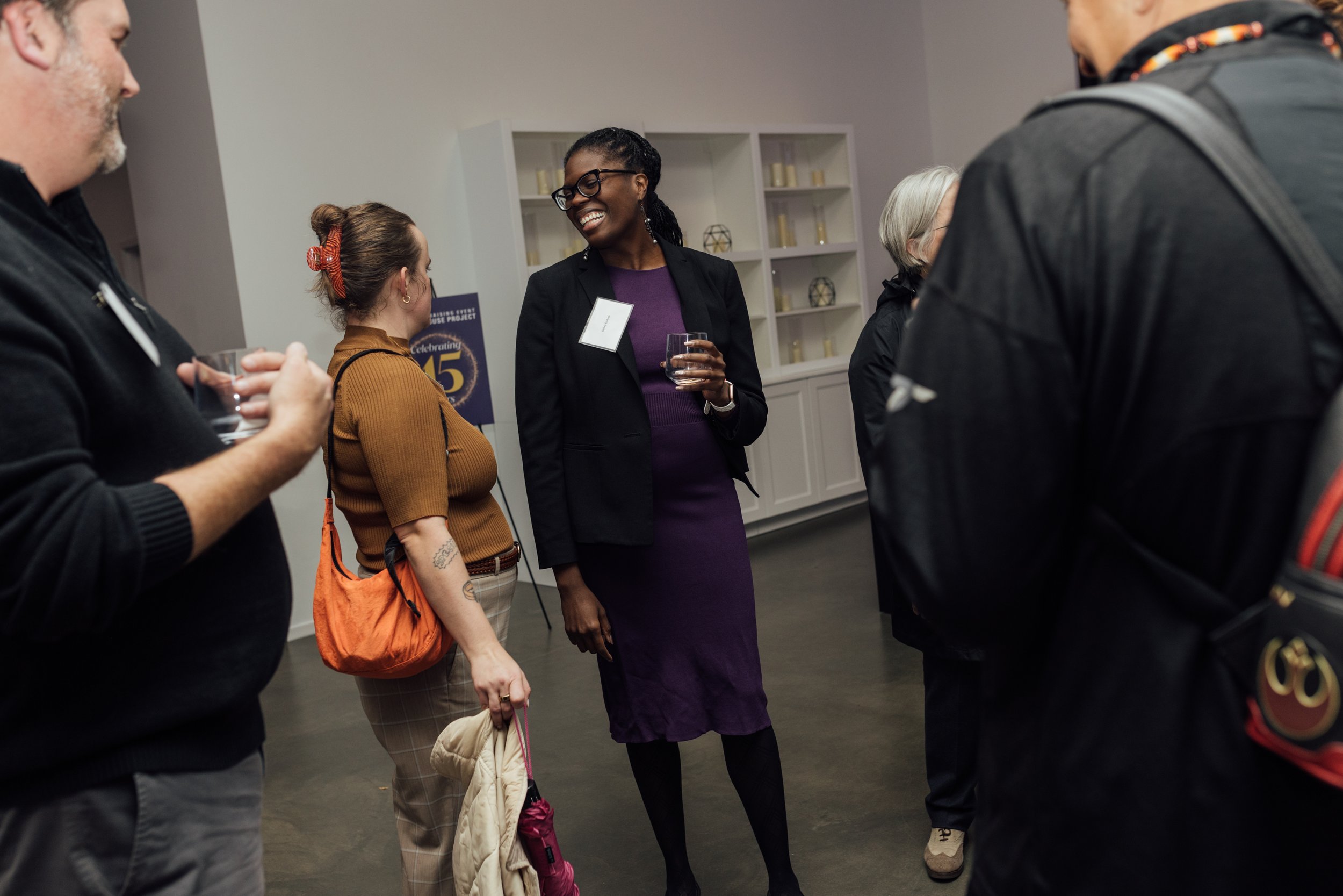 Group of diverse people at a social event, engaged in conversation, with a woman in a purple dress and black blazer smiling and holding a glass of water.