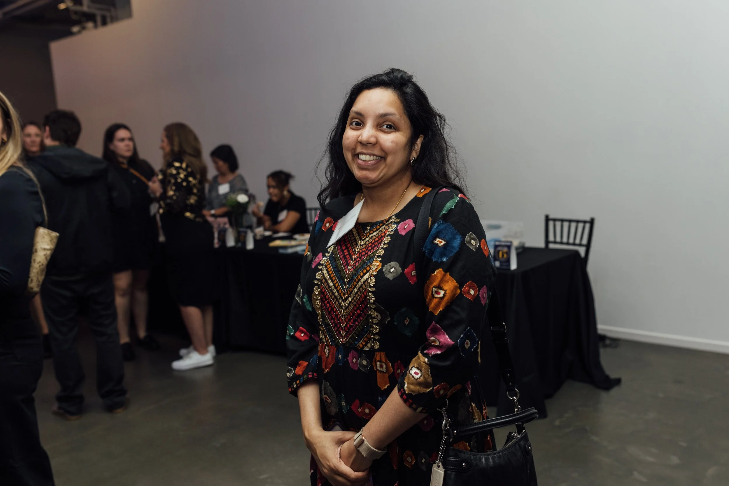 A woman smiling at an indoor event, with people and a table with items in the background.