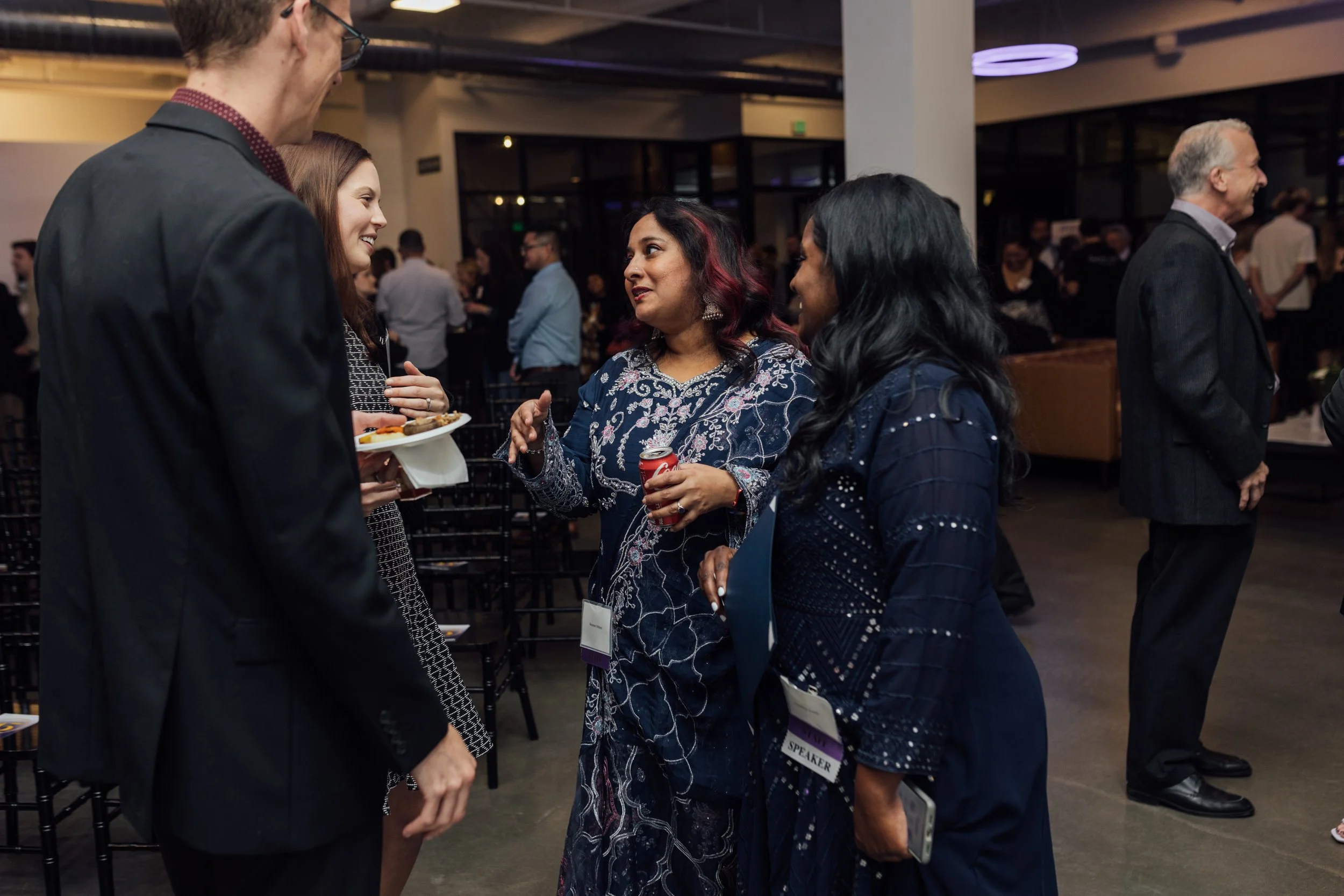 A group of four diverse people, three women and one man, engaged in conversation at a professional event, with some holding plates of food and drinks, in a spacious conference hall with other attendees in the background.