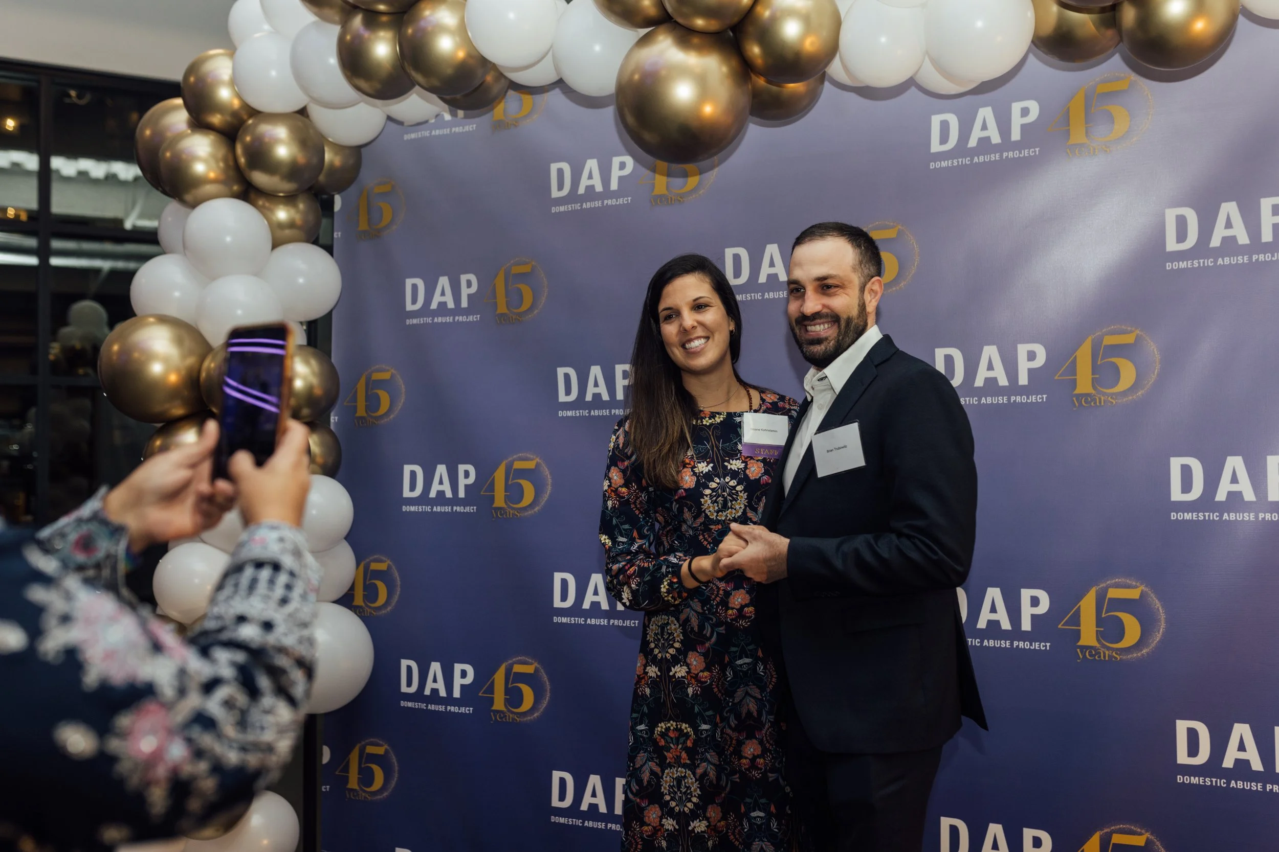 Two people, a woman and a man, are smiling and holding hands in front of a backdrop with the logo and text 'DAP 45 years'. The woman has long dark hair and is wearing a floral dress. There is a cluster of white and gold balloons above them, and a per