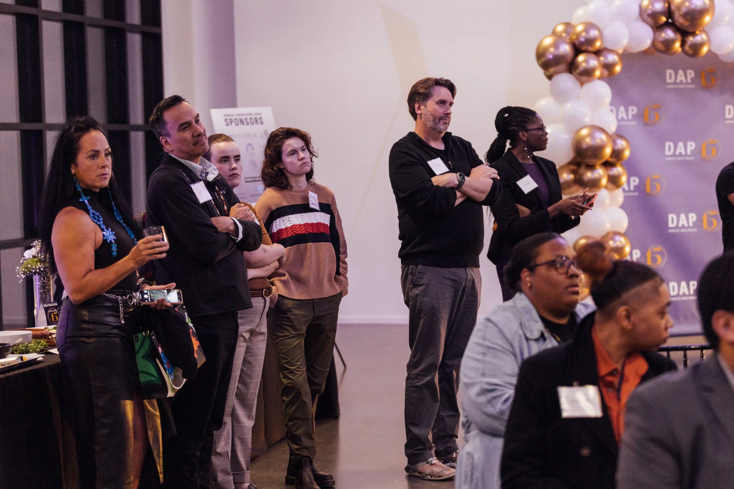 Group of diverse adults and children attending a formal event, standing and sitting, with a backdrop decorated with balloons and the text 'DAP 15'.