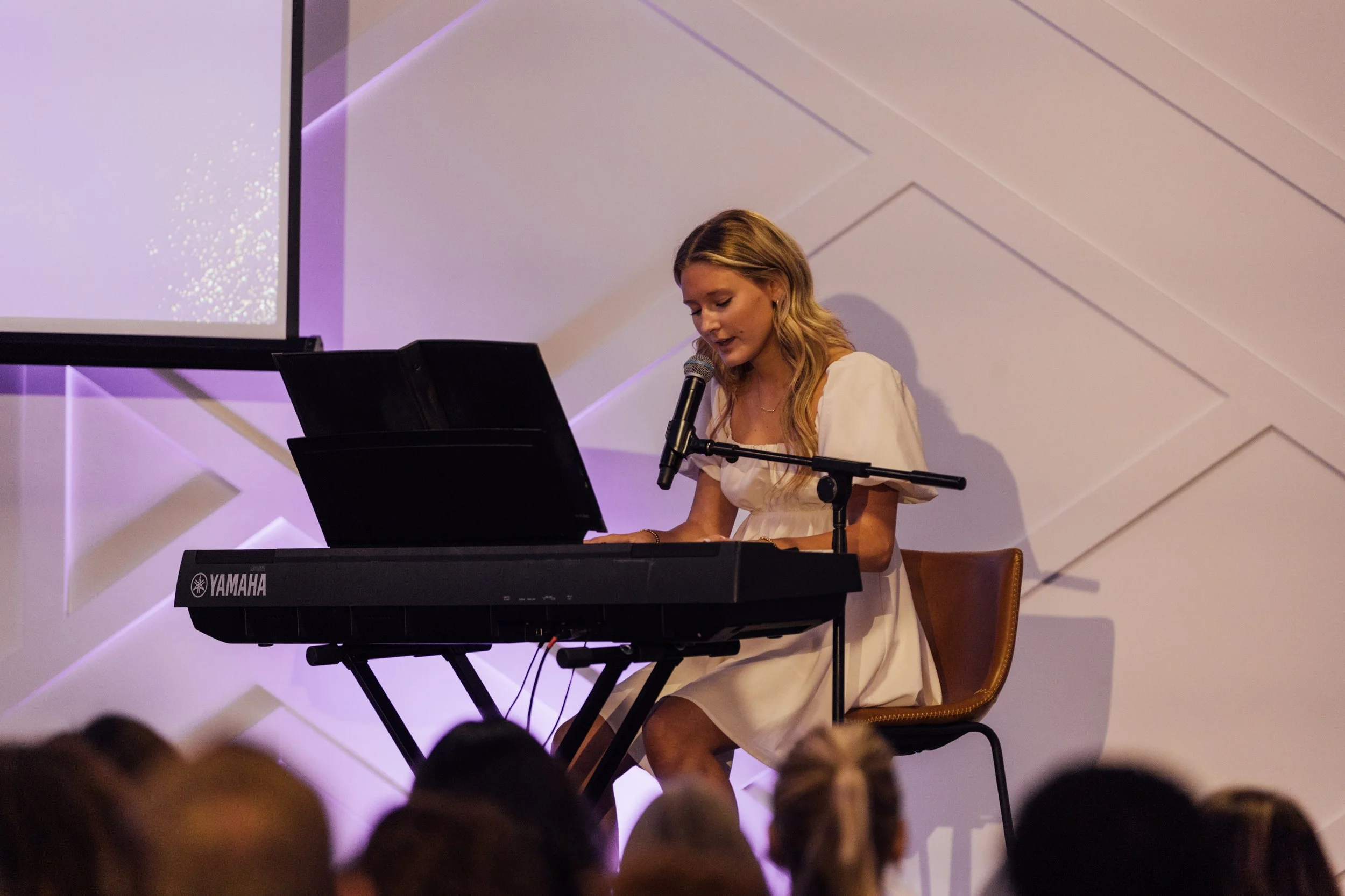 A young woman in a white dress plays a Yamaha keyboard and sings into a microphone on stage during a performance.