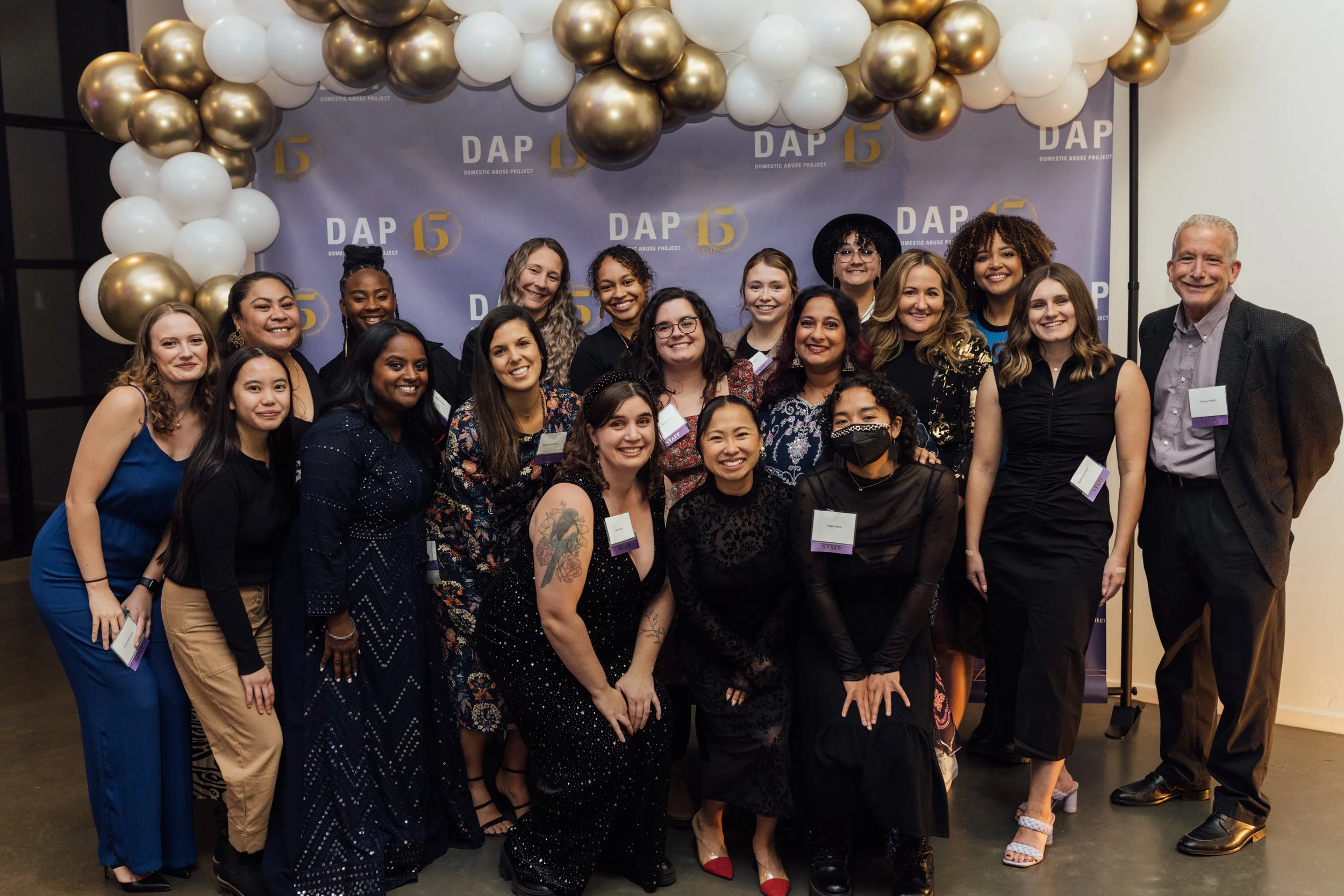 Group photo of diverse women and one man at a celebration event, standing in front of a backdrop with balloons and a logo that reads 'DAP 15' and 'Domestic Abuse Project', with most individuals smiling at the camera.