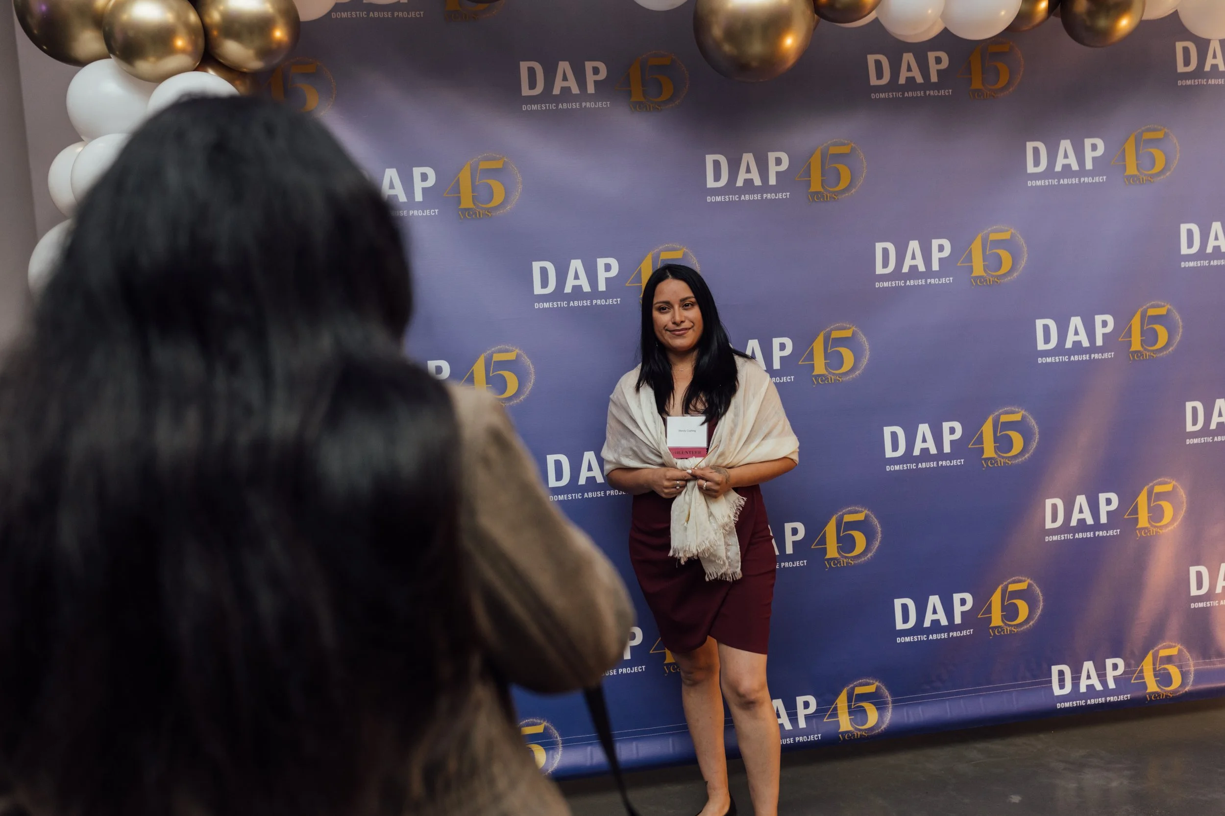 A woman poses in front of a blue backdrop with the logo and text for the Domestic Abuse Project's 45th anniversary celebration. There are gold and white balloons on the top left corner. The woman is smiling, wearing a dark dress, and has a scarf arou