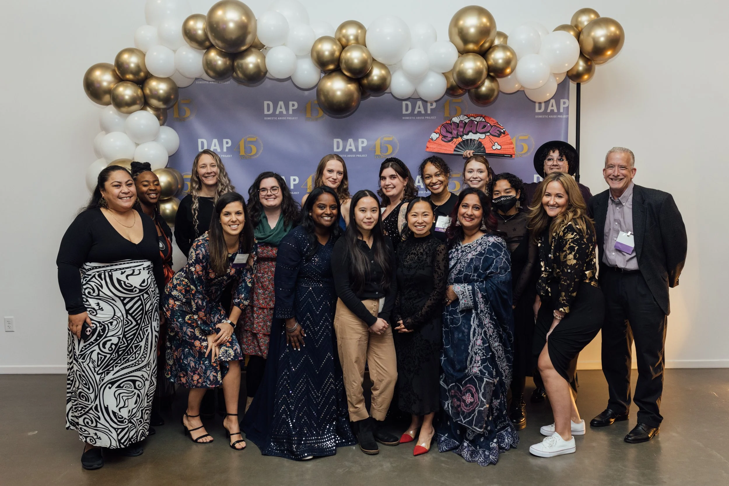Group of diverse women and men smiling at an event with a backdrop that says DAP 45, decorated with white, black, and gold balloons
