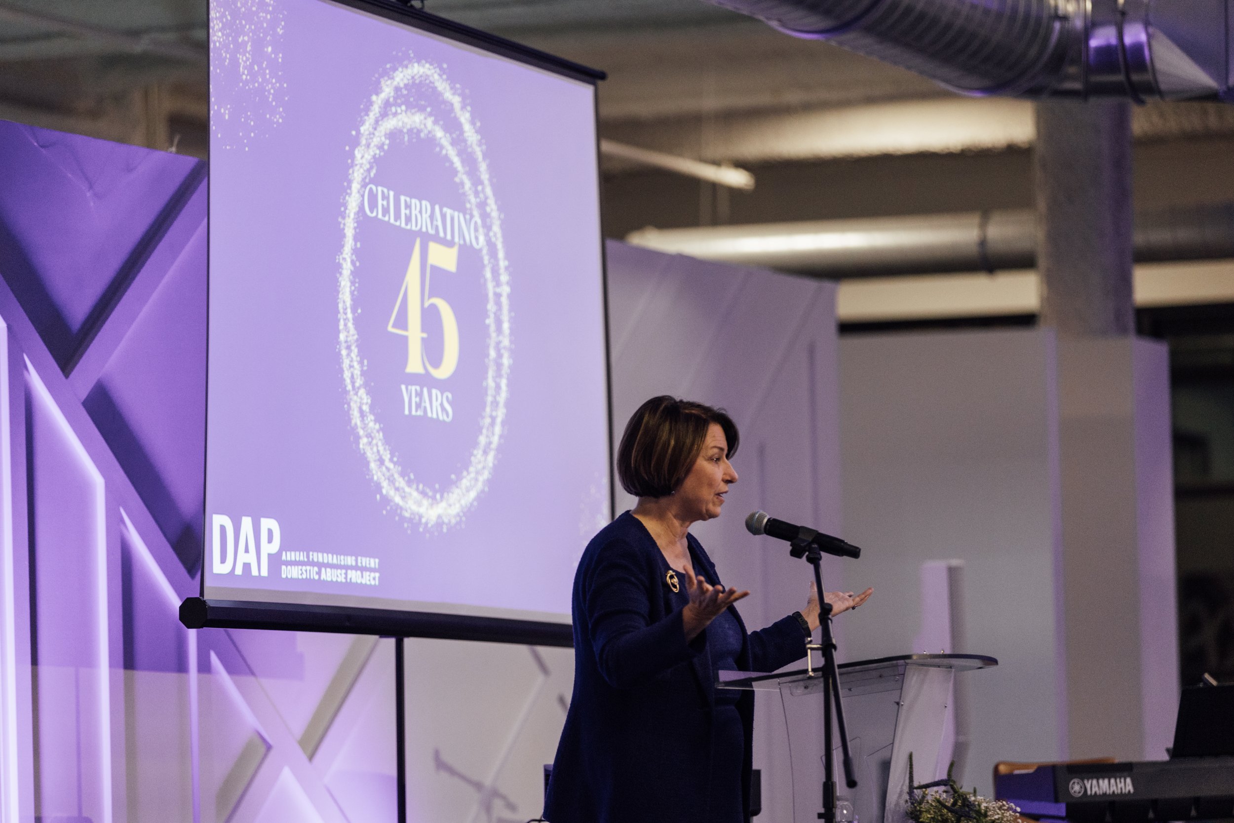 A woman giving a speech at an event celebrating 45 years, with a large screen behind her displaying the same message, in a decorated indoor space.