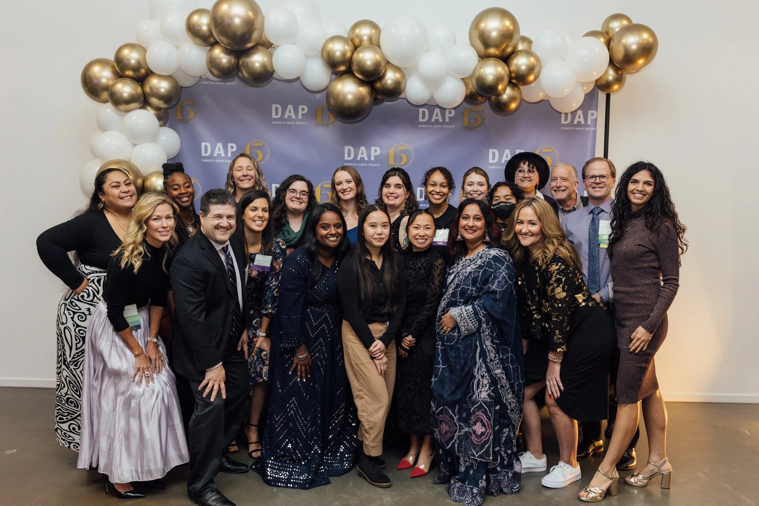 Group of diverse people dressed in formal and semi-formal attire posing for a group photo at an event with a backdrop that says DAP 15 and balloons in gold, white, and black.