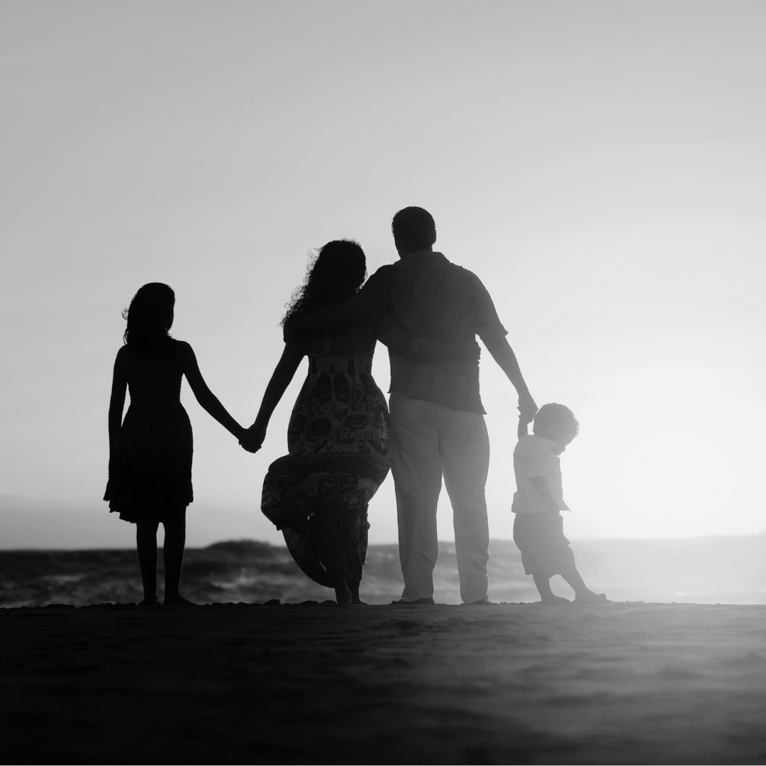 Silhouettes of a family holding hands on the beach during sunset, with two adults and two children, in black and white.