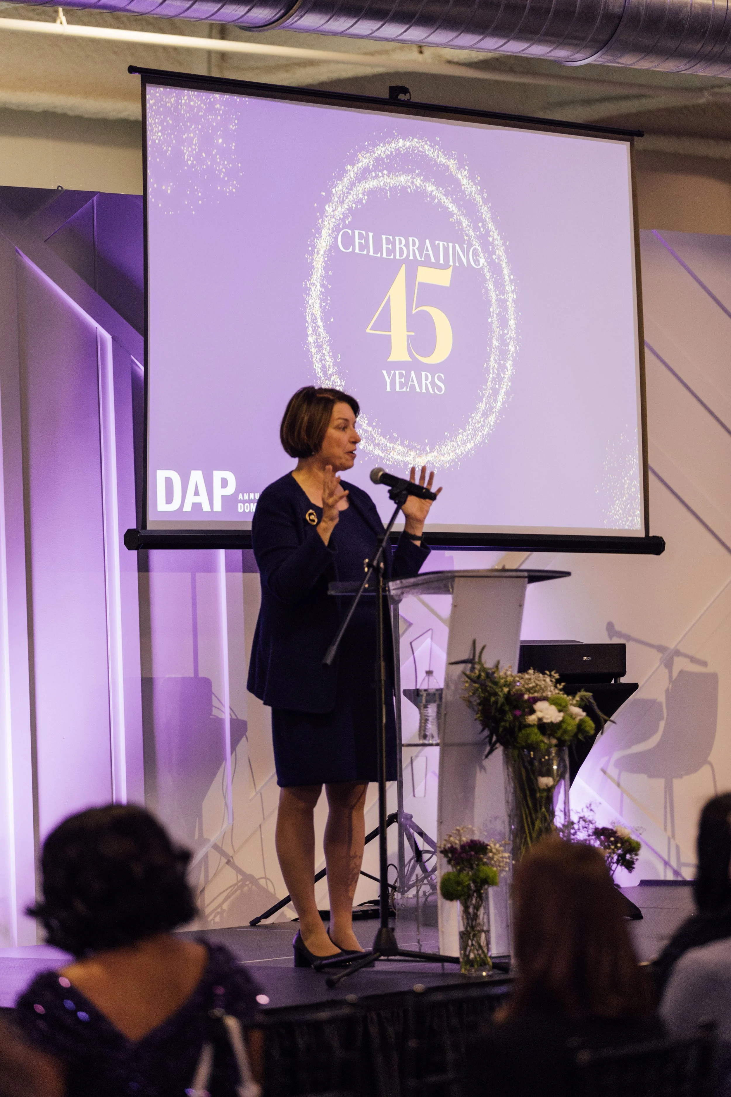 A woman in a dark suit standing at a podium, speaking with a microphone, at an event celebrating the 45th anniversary of DAP, with a large screen behind her displaying 'Celebrating 45 Years' and purple decorations.