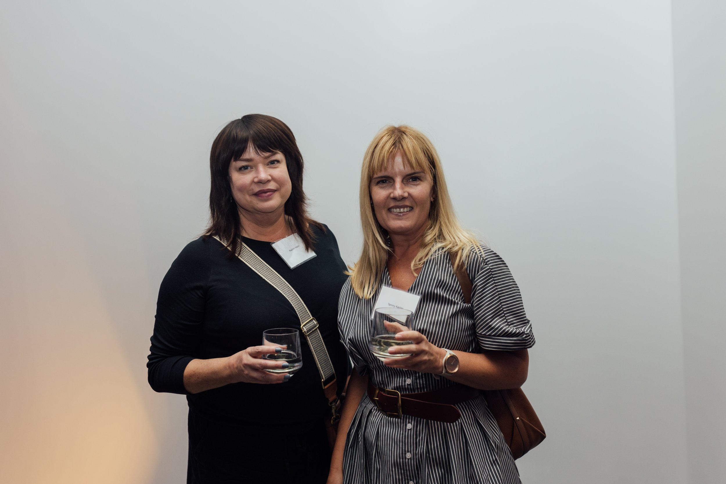 Two women with name tags are standing together, holding glasses of water at an indoor event.