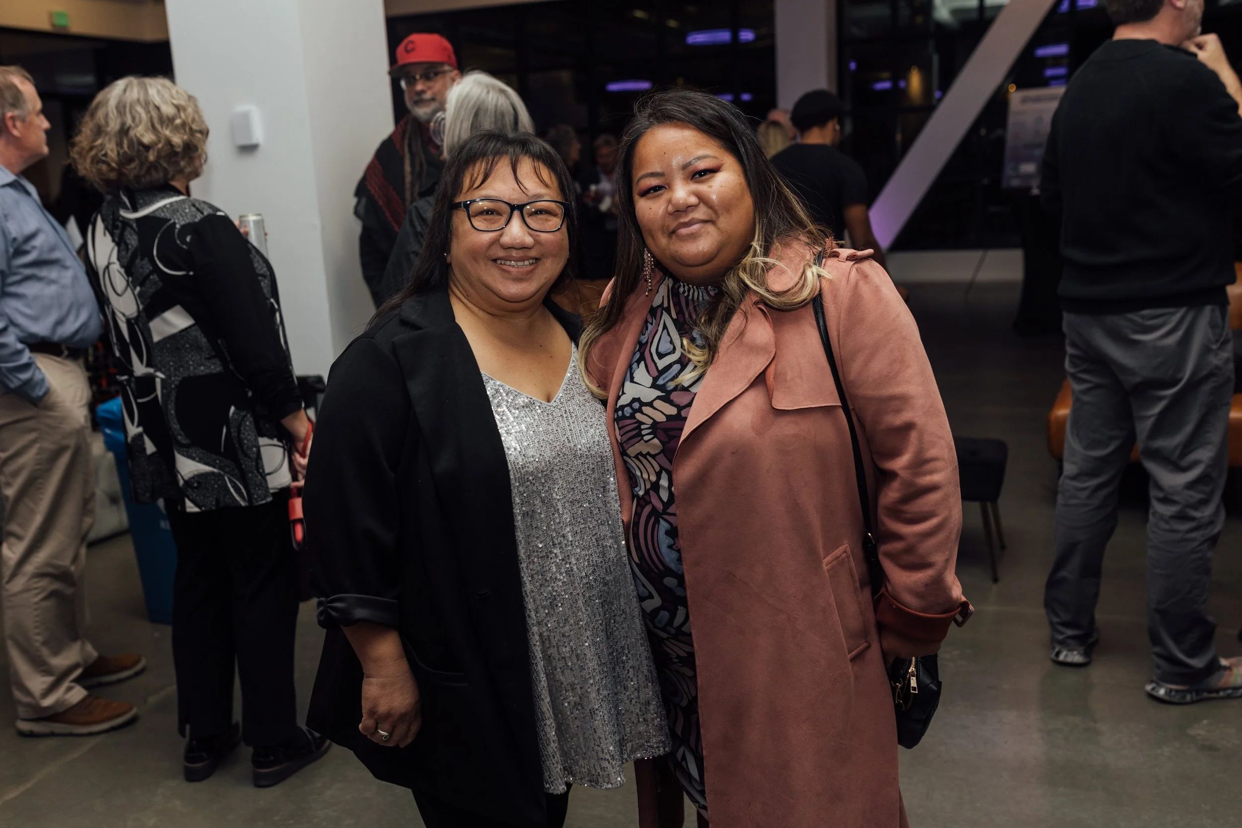 Two women smiling and posing for a photo in a crowded indoor event space.