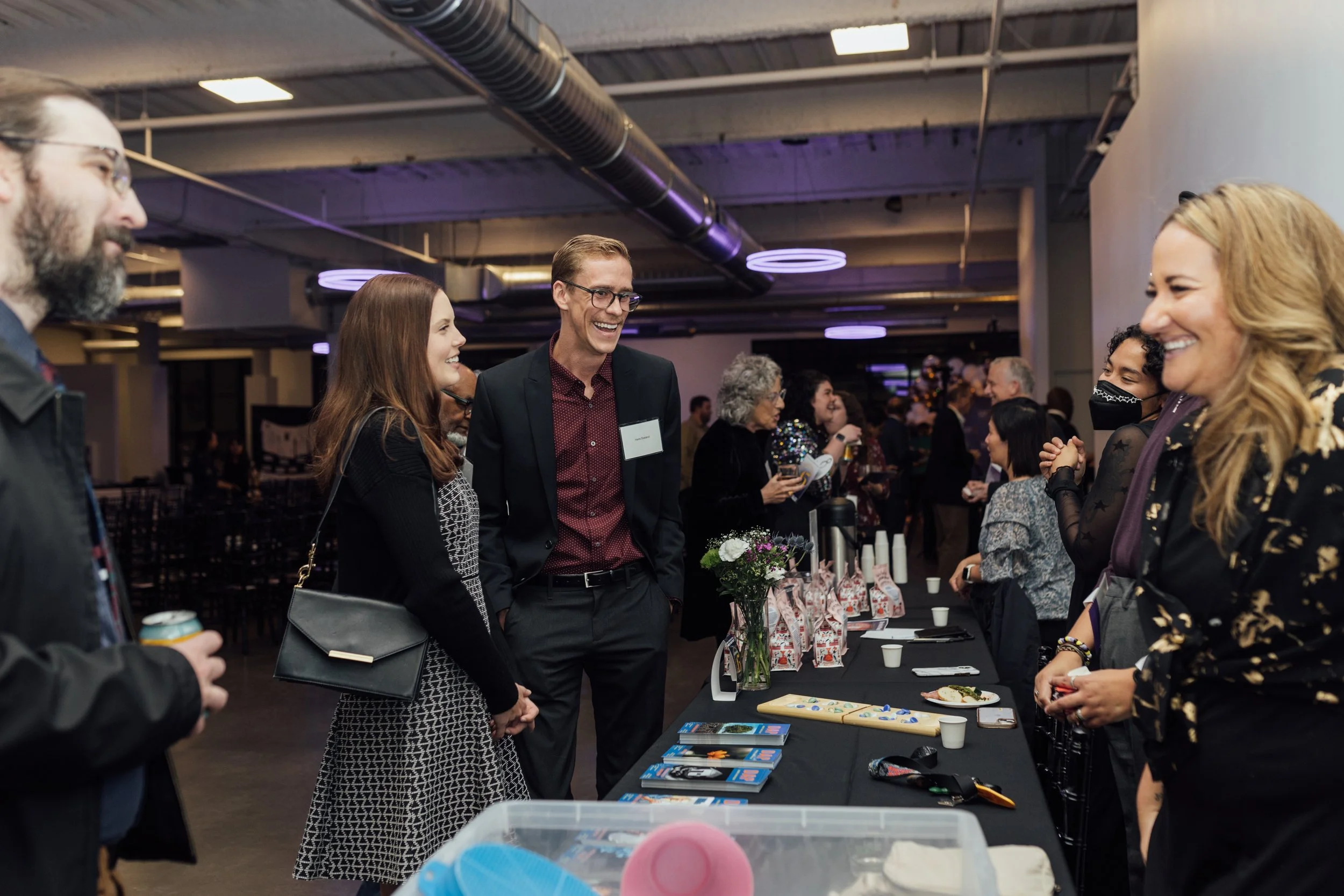 People smiling and chatting at a social event or reception with tables displaying snacks and brochures in a modern indoor venue