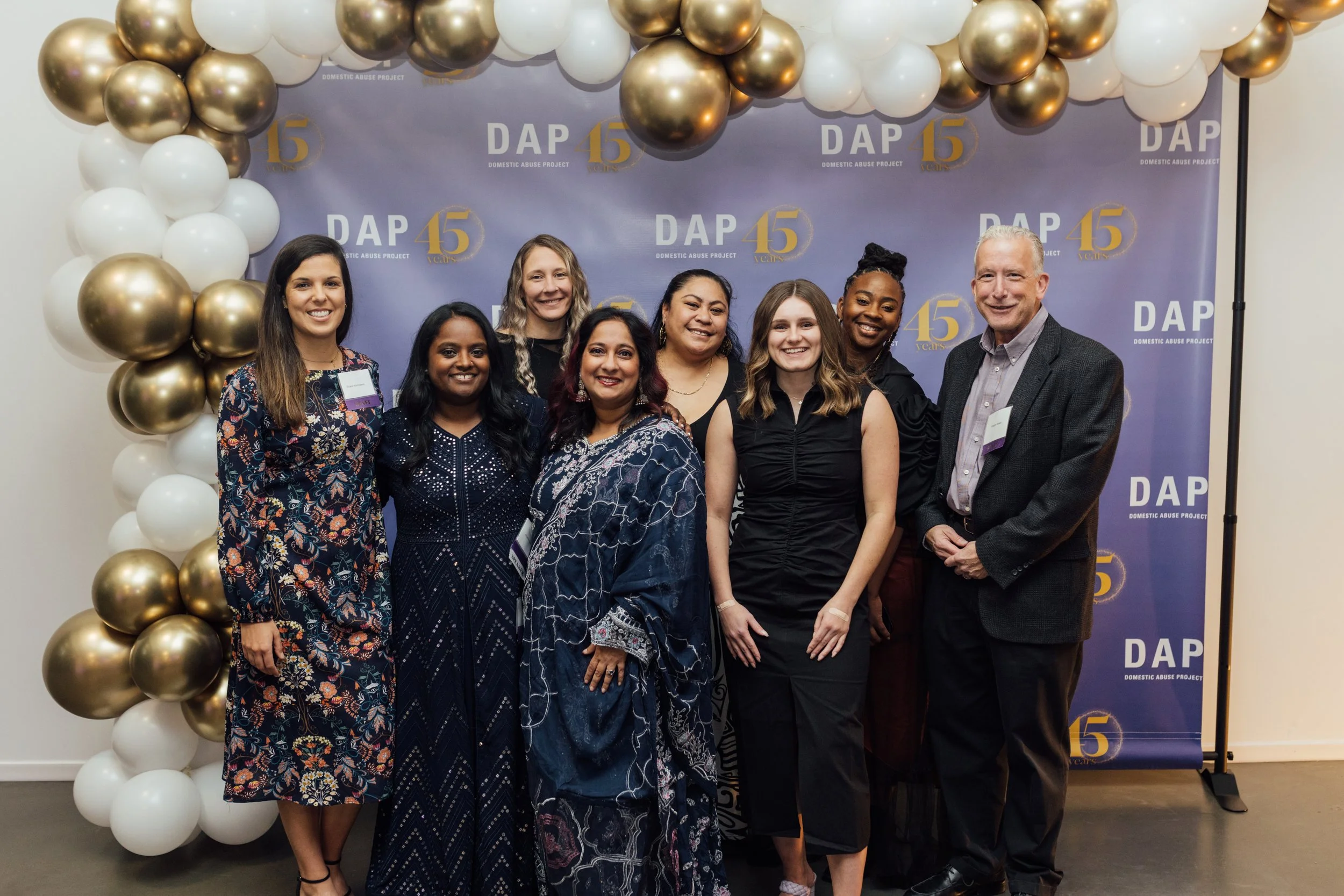 Group of eight people standing together in front of a purple backdrop with the logo for Domestic Abuse Project's 45th anniversary, decorated with white, gold, and black balloons.