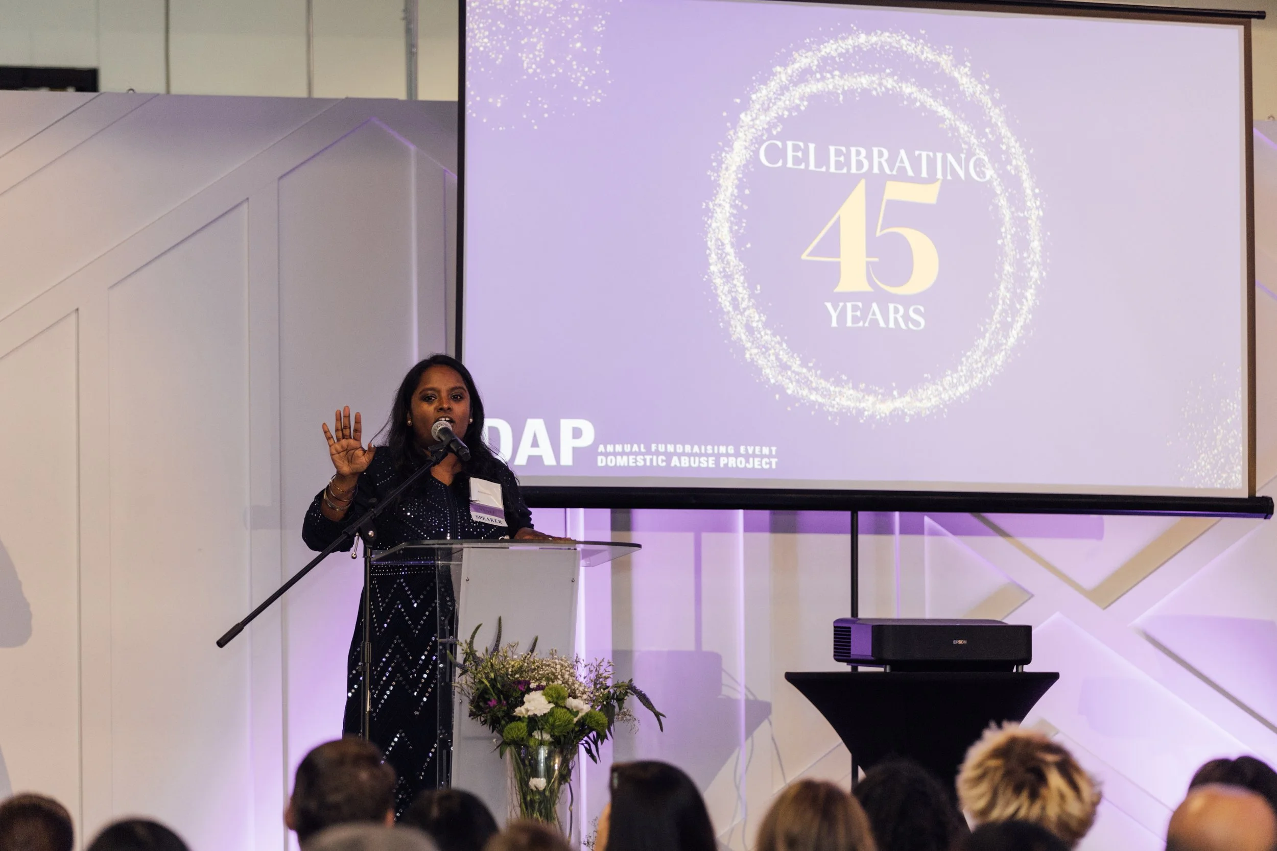 Woman speaking at podium during a 45th anniversary celebration event with a large screen displaying 'Celebrating 45 Years' in the background.