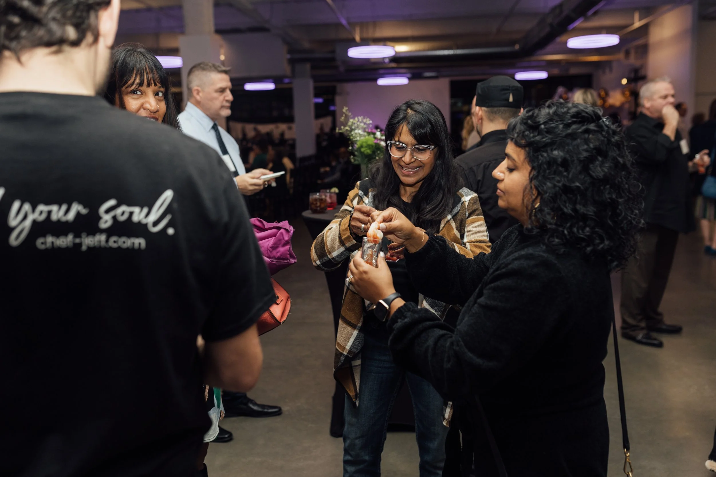 People socializing at an indoor event, with two women in the center handling a small item, surrounded by other guests.