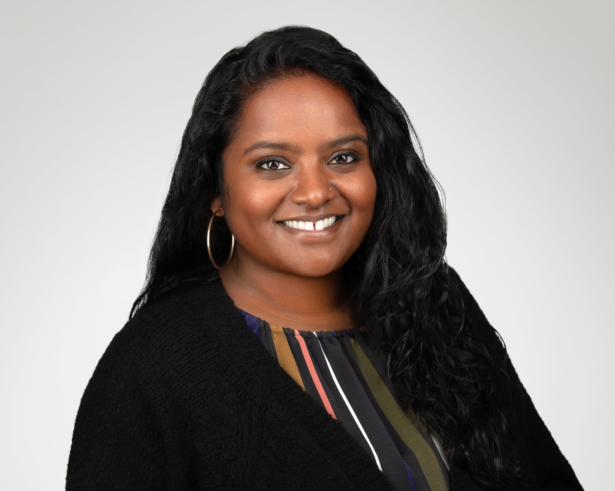 A woman with dark, wavy hair wearing a black cardigan and hoop earrings, smiling against a plain light gray background.