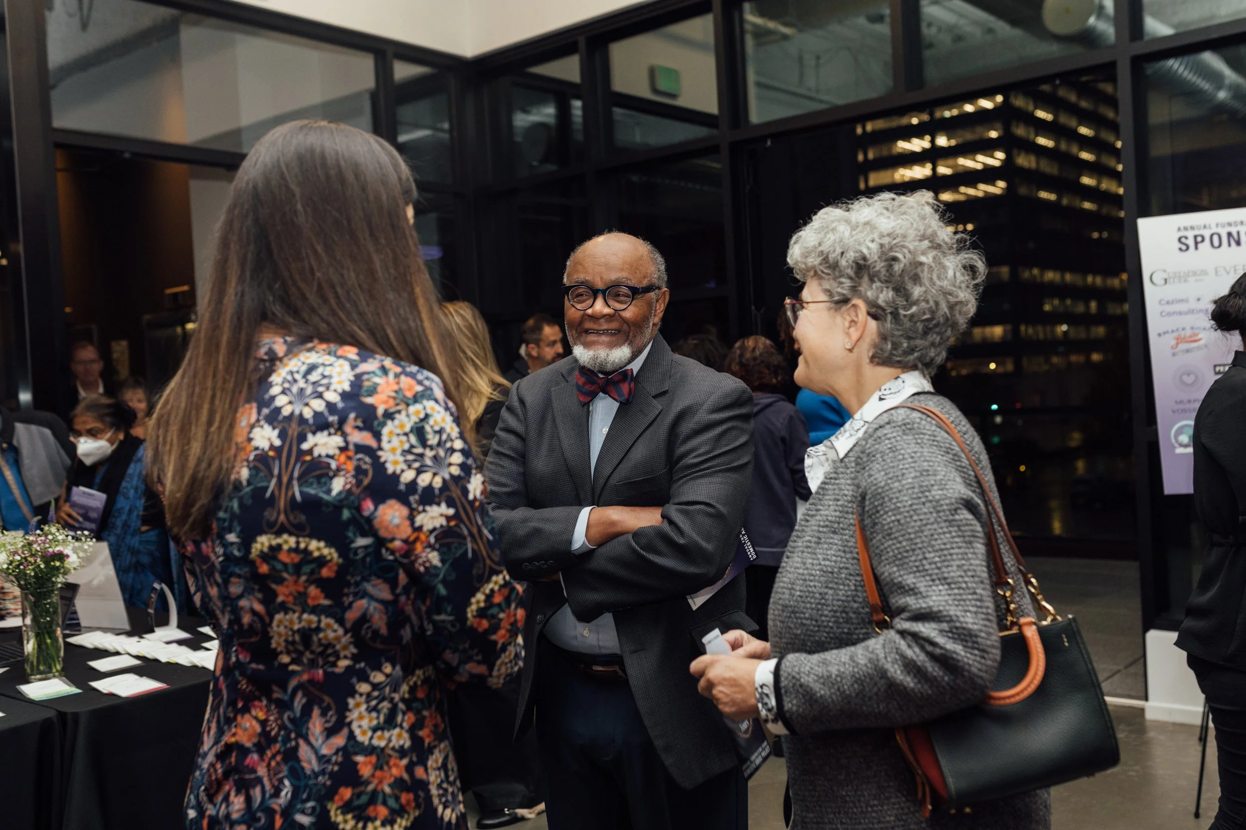 People at a networking event engaging in conversation, with large glass windows and a cityscape in the background.