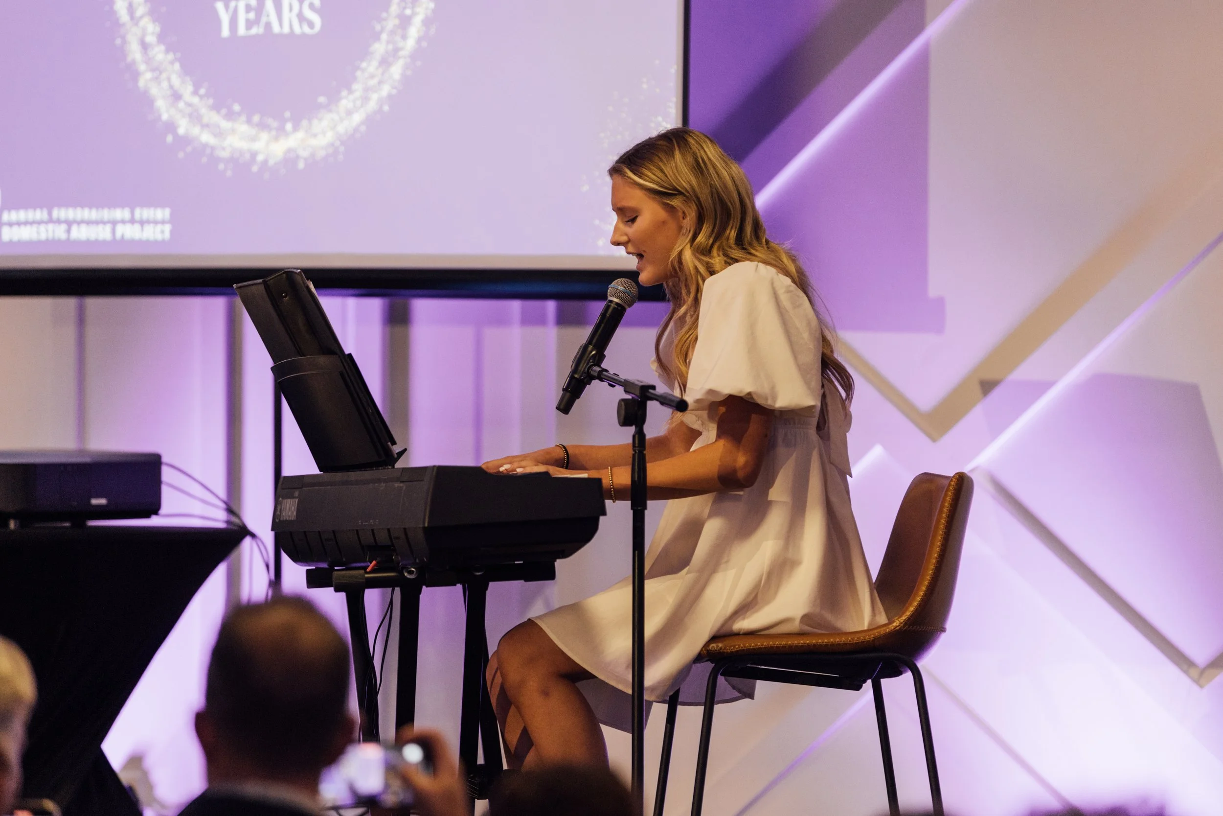 A woman singing and playing the keyboard at an event, with a microphone in front of her and a large screen behind her displaying part of an award or recognition event.