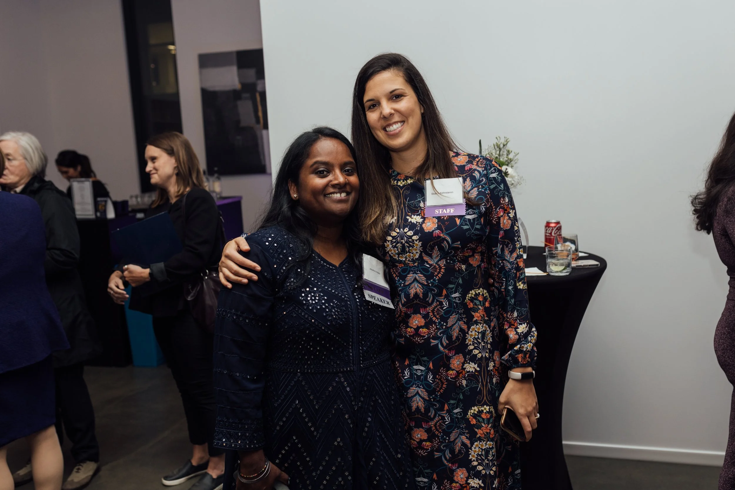 Two women smiling at a professional event, one with a 'SPEAKER' badge and the other with a 'STAFF' badge, standing close together in front of a plain white wall.