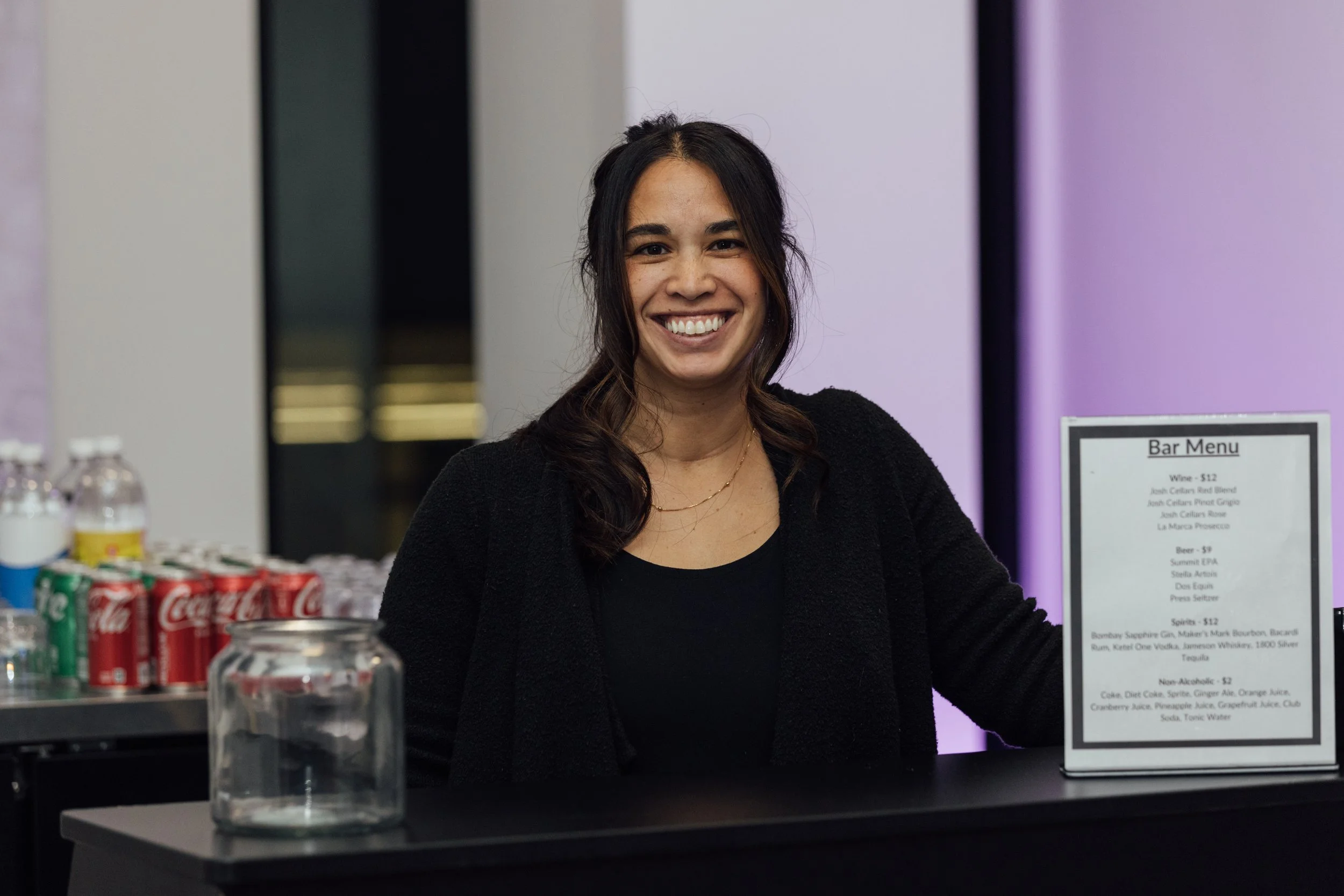 A smiling woman standing behind a bar counter with soda cans and bottles on the side and a bar menu on the right.
