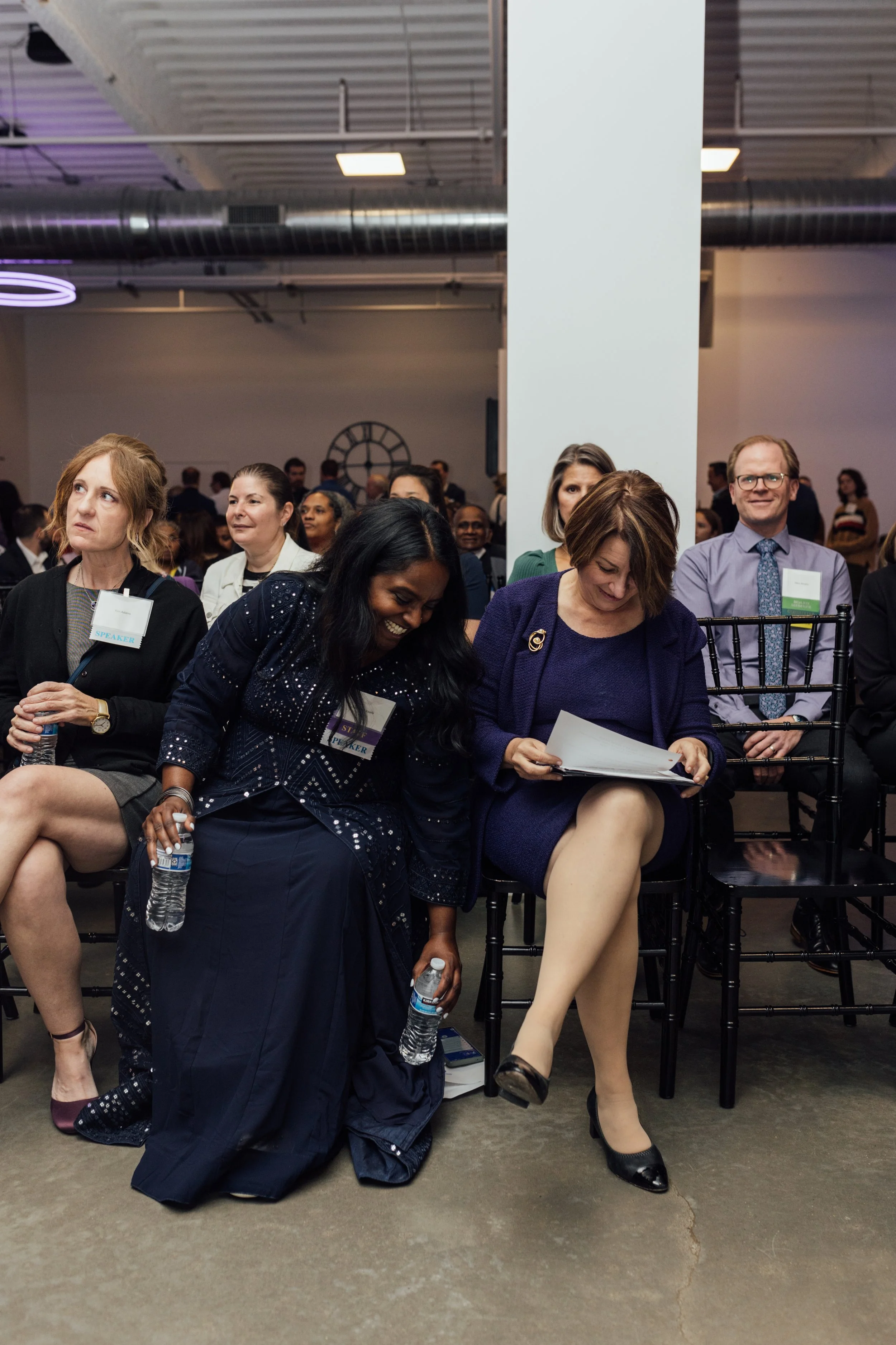 A group of people sitting in an auditorium or conference room, some holding water bottles, with a mix of men and women, in professional attire, attending an event or conference.