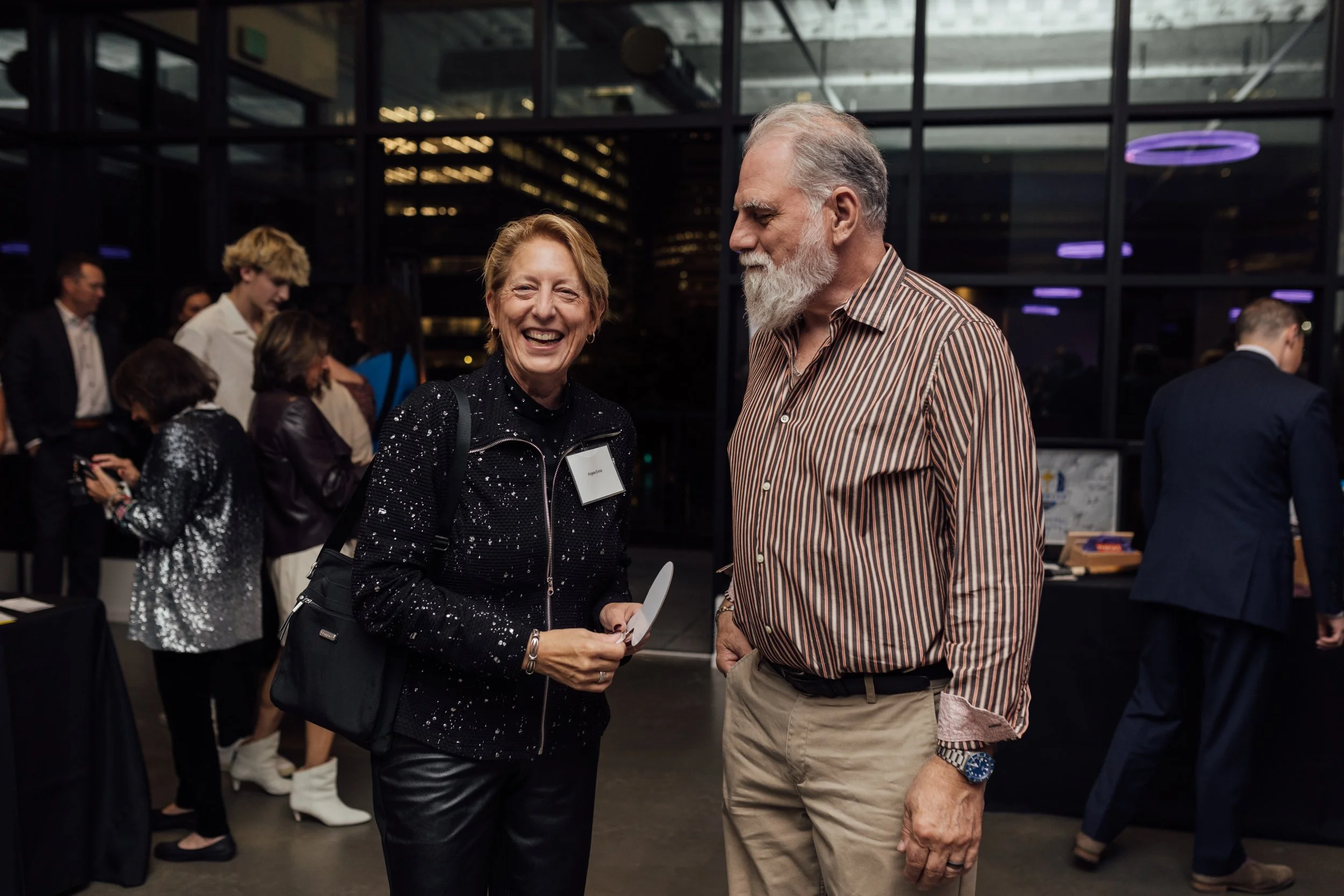 Two elderly people smiling and talking at a social event with other attendees in the background.