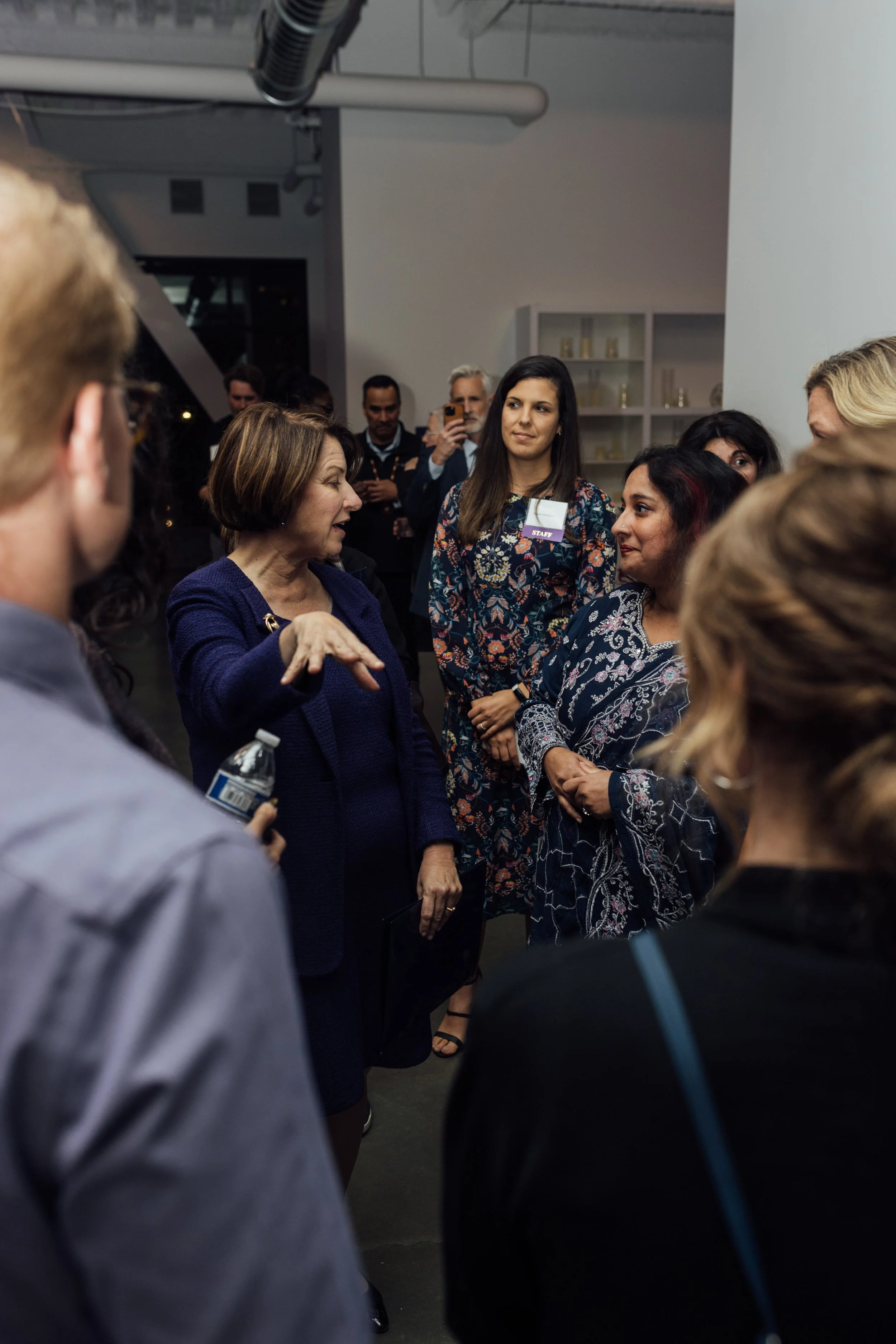 A group of people talking at an indoor event, with one woman in a blue blazer speaking to a diverse group of attendees.