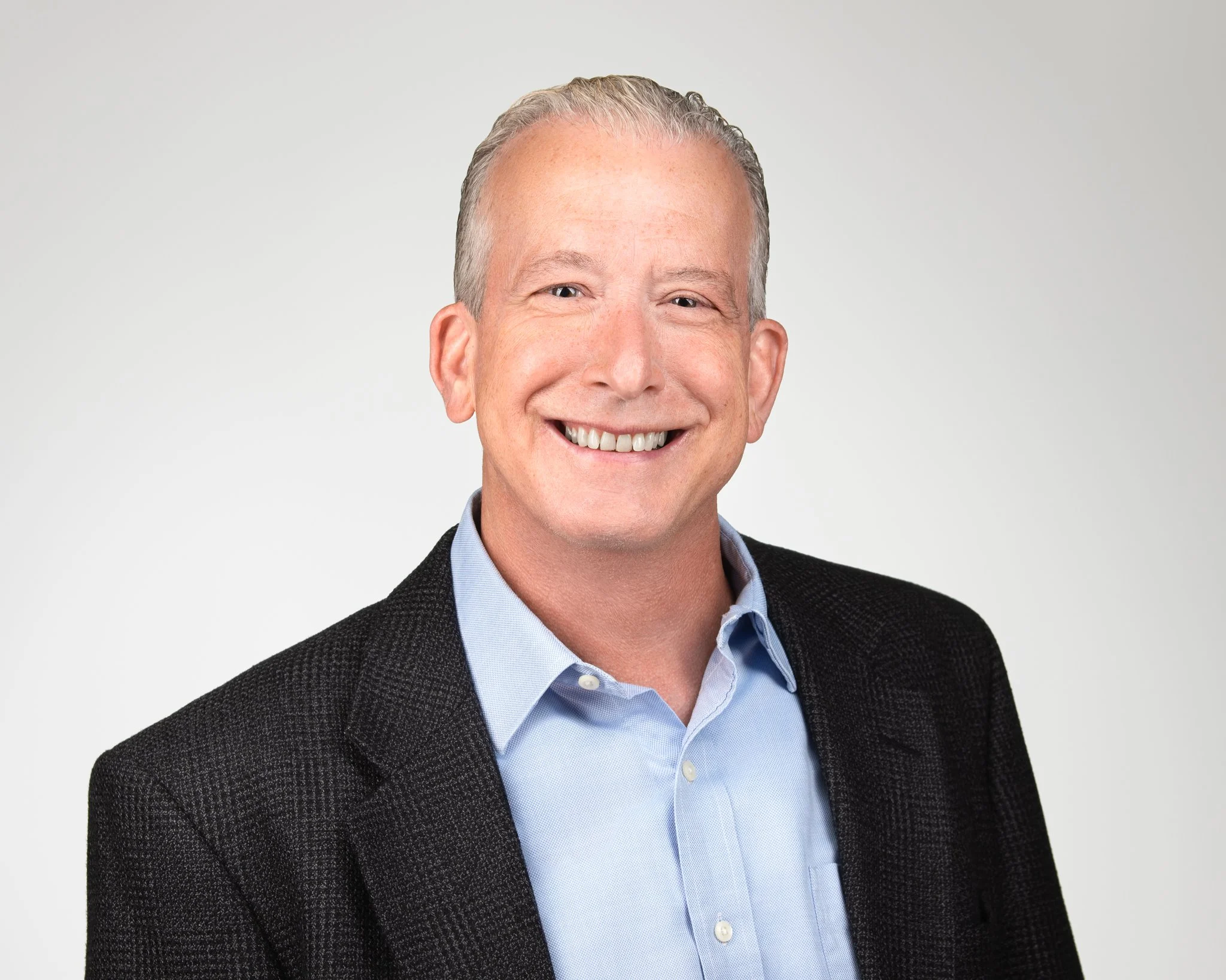 Headshot of a middle-aged man with gray hair, smiling, wearing a light blue collared shirt and black blazer, against a plain gray background.