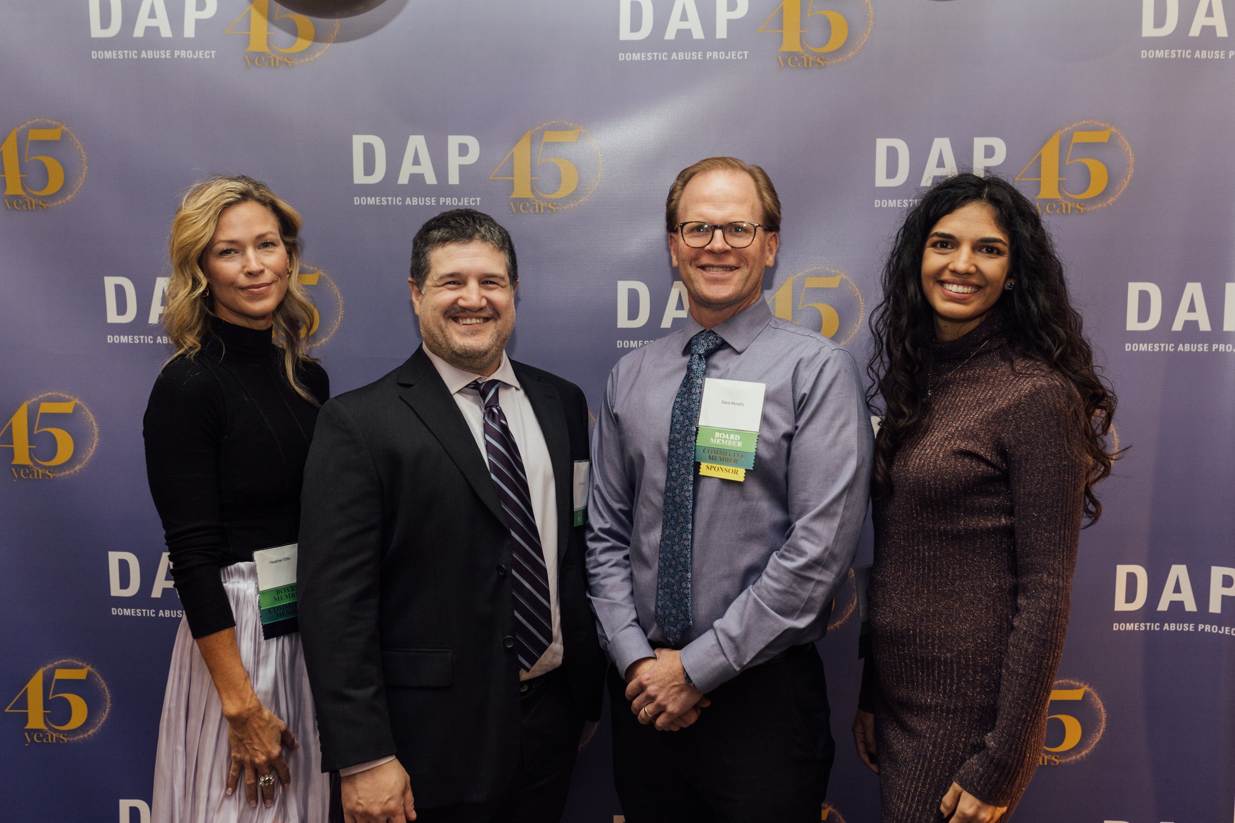 Four people standing in front of a backdrop with the DAP 45 logo, attending a Domestic Abuse Project event. They are dressed in formal attire and all wearing name badges, smiling at the camera.