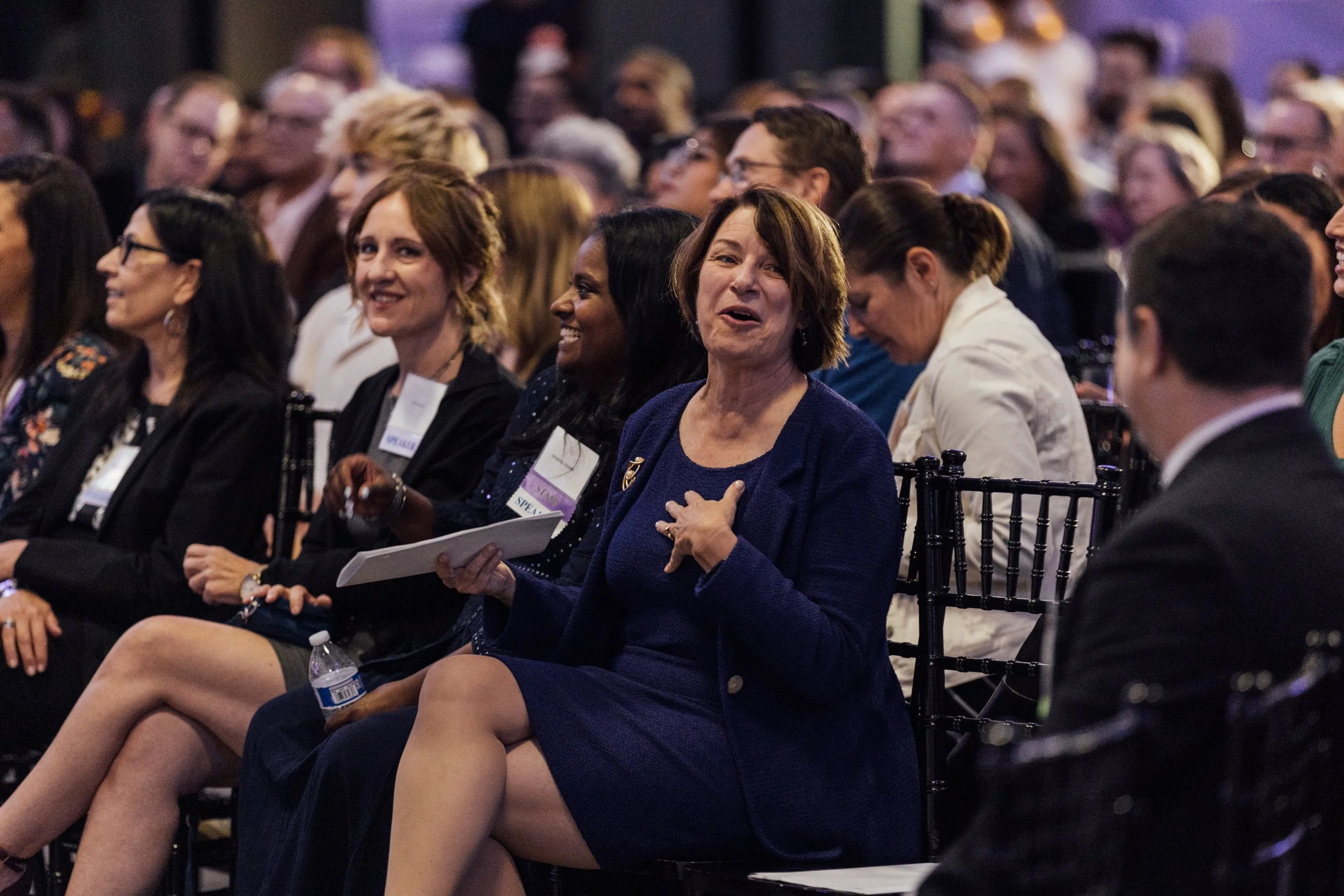 A woman in a blue blazer and dress on stage appears emotional, clutching her chest and holding a notebook, surrounded by an audience in a conference setting.