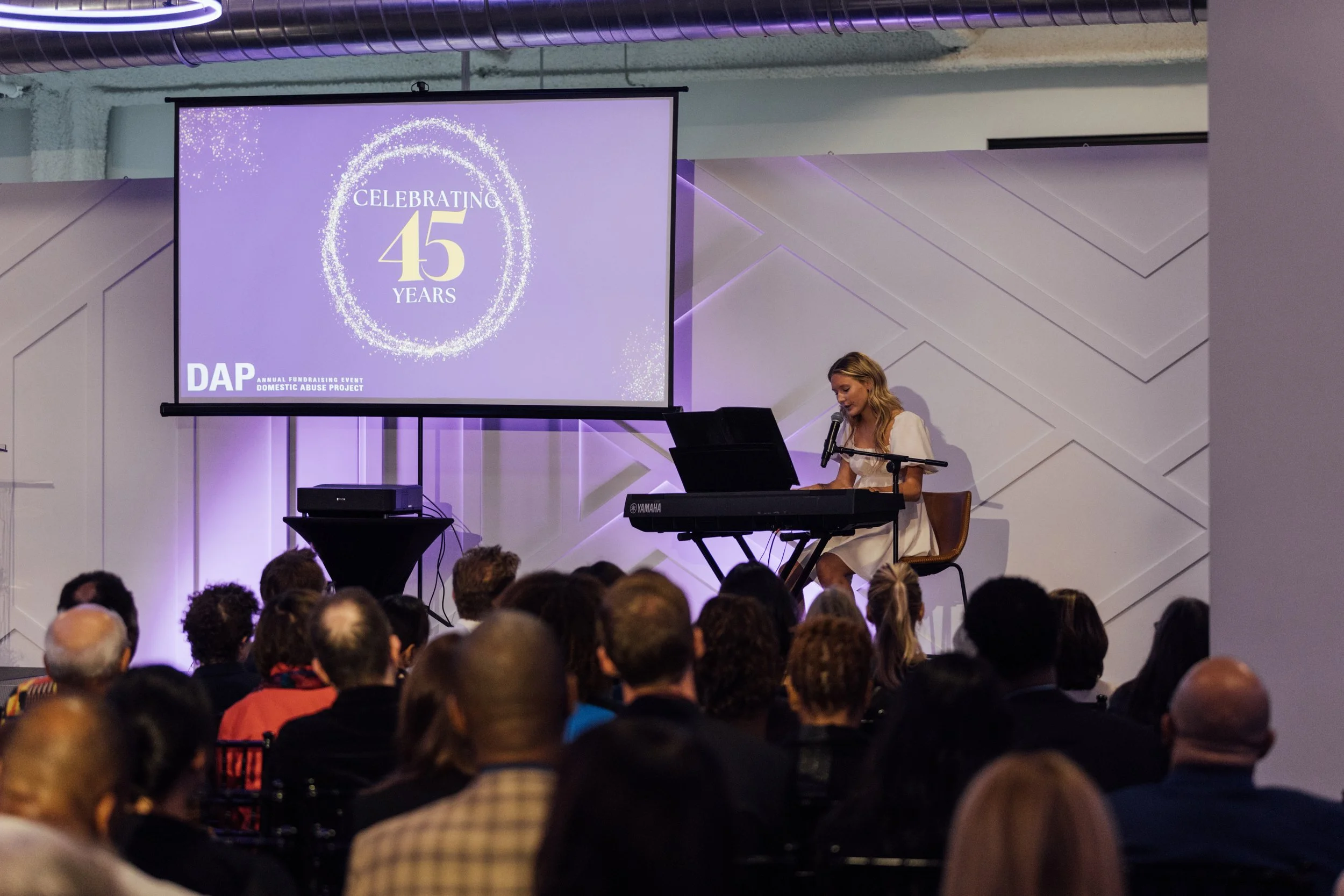 A woman in a white dress playing a keyboard and singing into a microphone at a stage during a celebration event. The screen behind her displays a graphic with the text 'Celebrating 45 Years' and the logo for DAP, an organization supporting domestic a