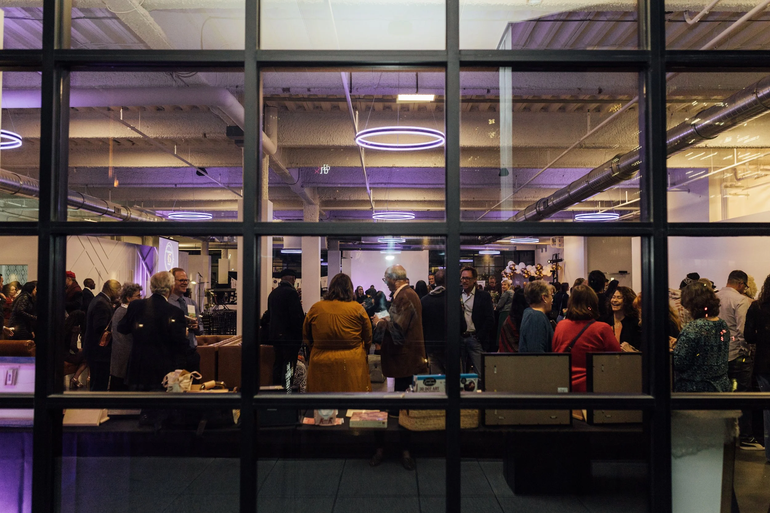 People gathered in an indoor event space with purple lighting and modern circular ceiling lights, seen through a large window with a grid pattern.