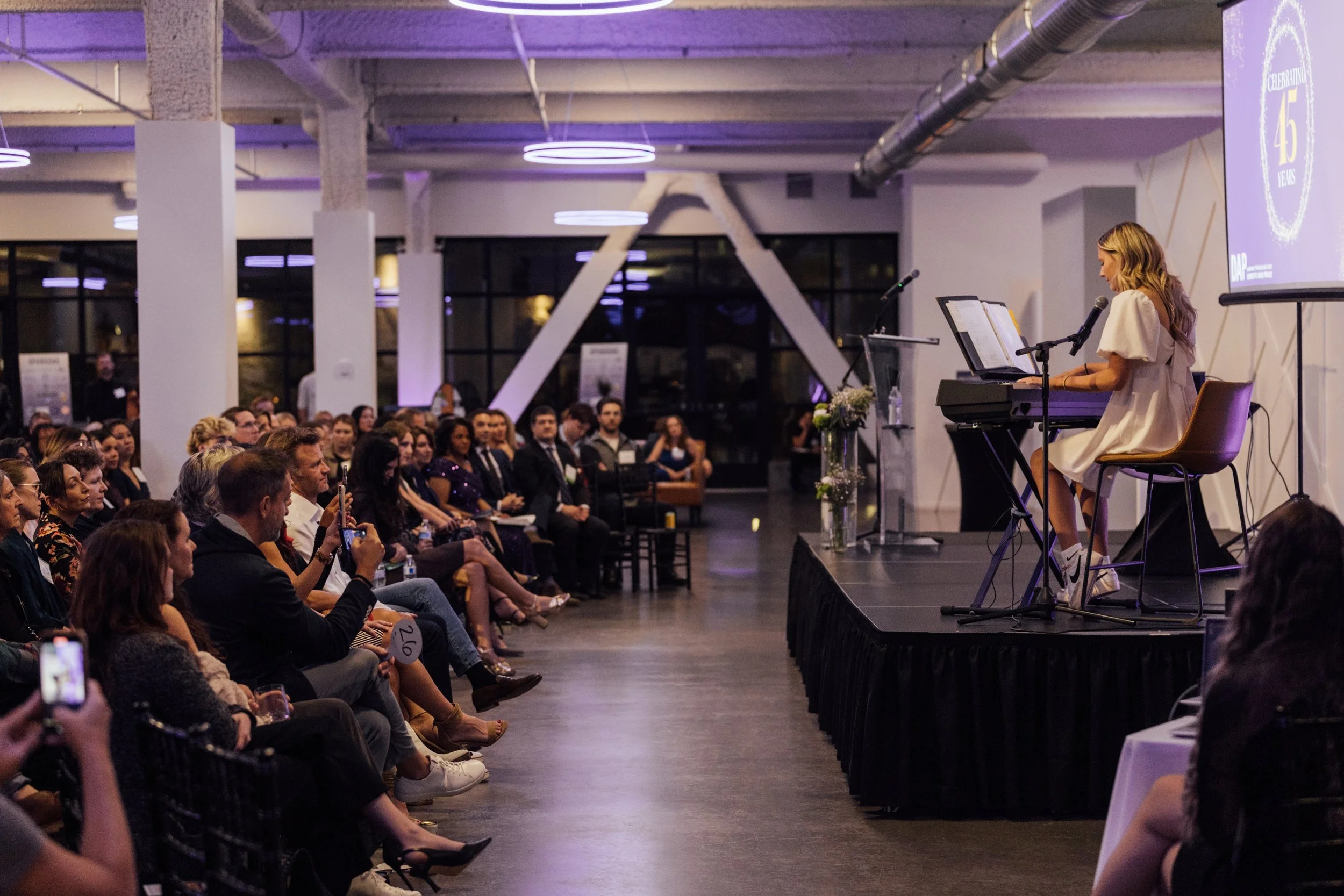 A woman dressed in a white dress performs on stage with a keyboard and microphone in front of a large audience at a formal event in a modern industrial-style venue.