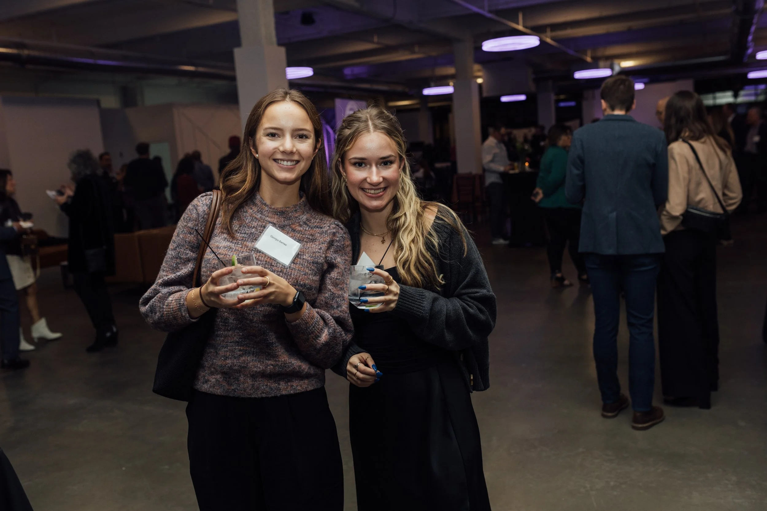 Two young women smiling at a social event, holding drinks, with other people mingling in the background under purple lighting.