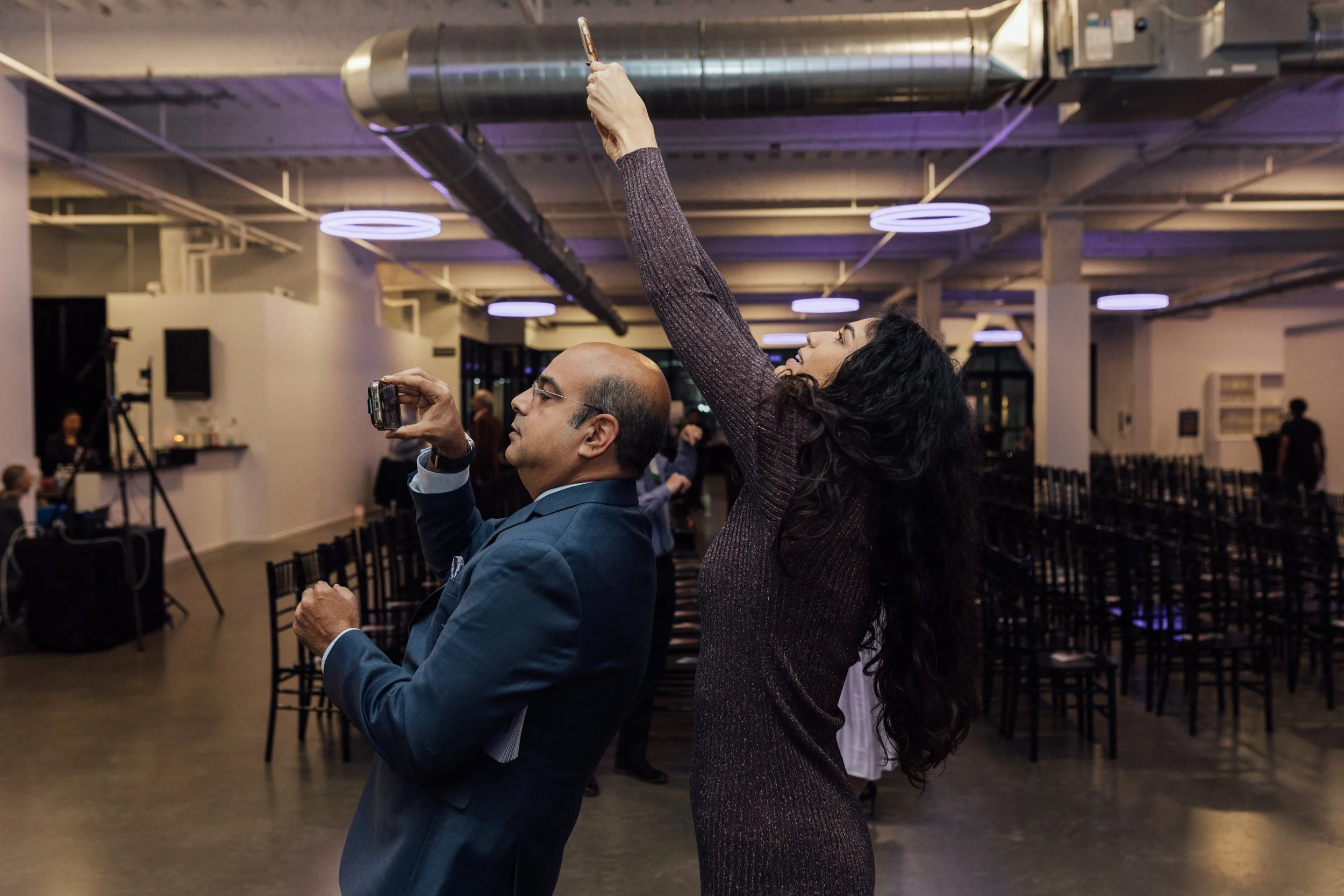 A woman with long dark curly hair, wearing a shimmering brown dress, is holding a smartphone above her head, taking a photo or video at an indoor event. A man in a blue suit is standing beside her, looking at her and holding a small camera or phone w